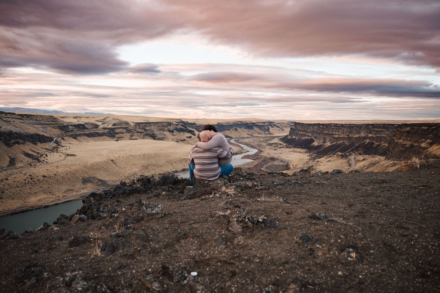 Couple standing on rocky cliff overlooking scenic river canyon during dramatic sunset.