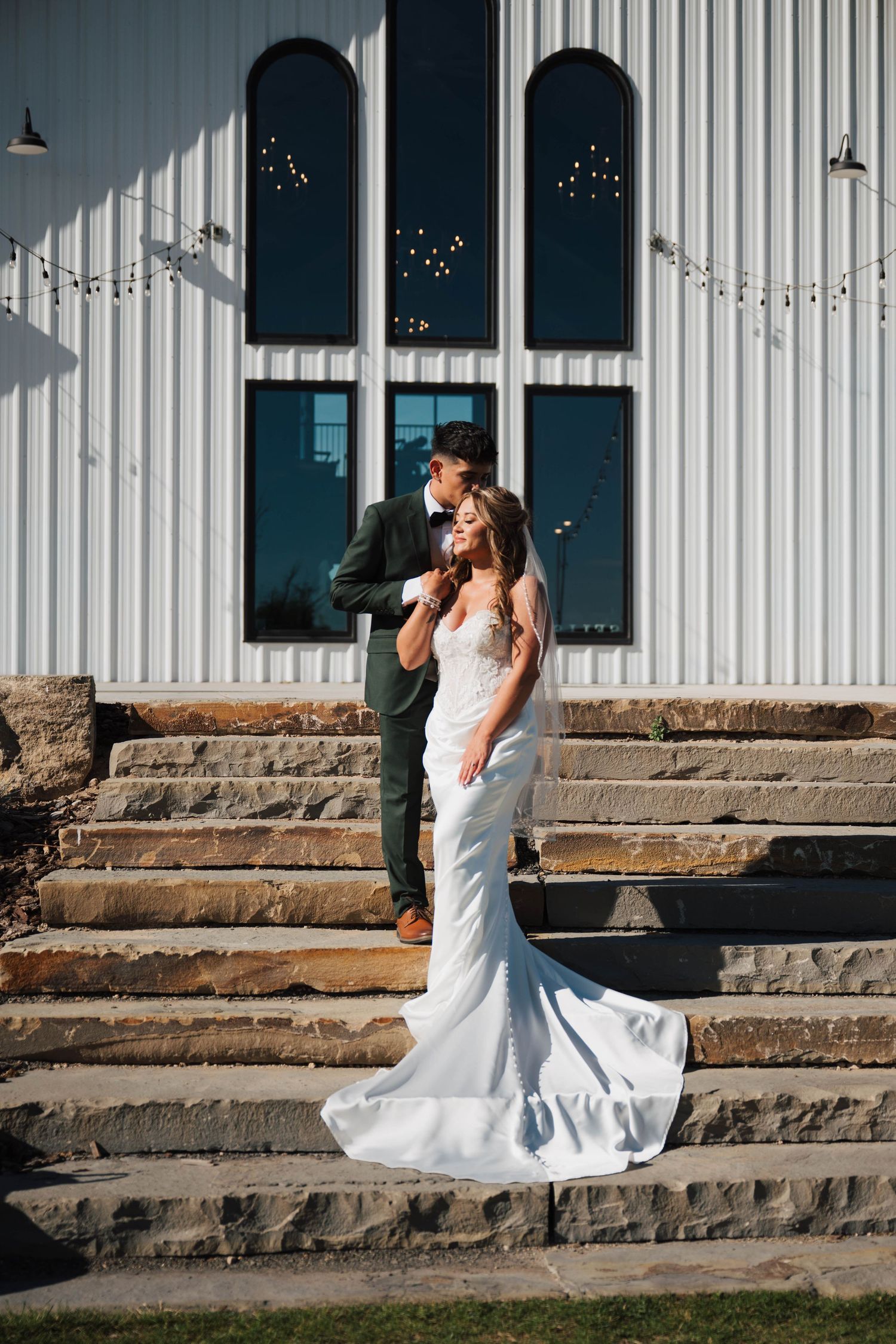 Bride in white gown and groom in dark suit pose on stone steps outside a white barn wedding venue.