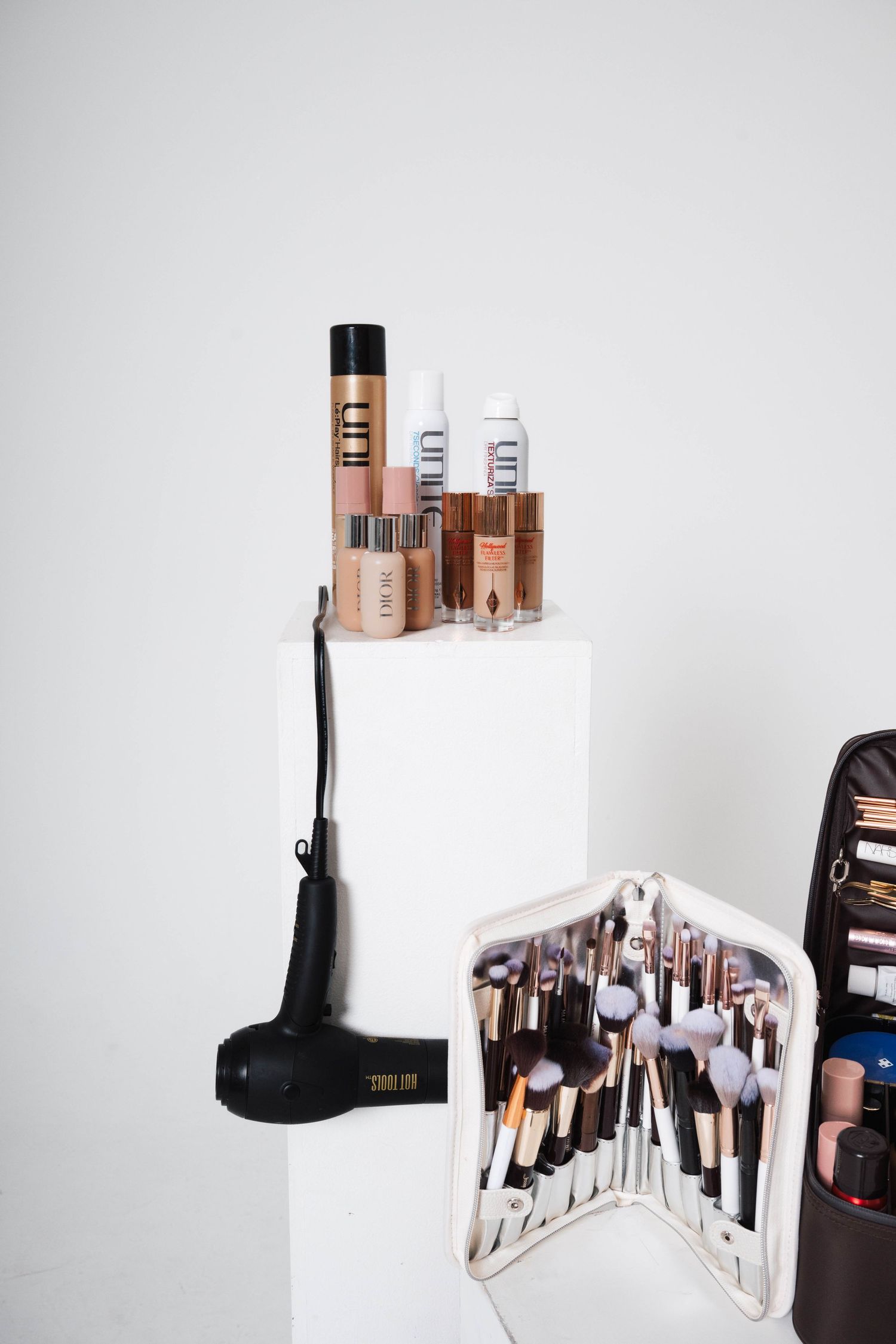 White pedestal displaying luxury makeup products, brushes in open case, and black hair dryer against white wall.