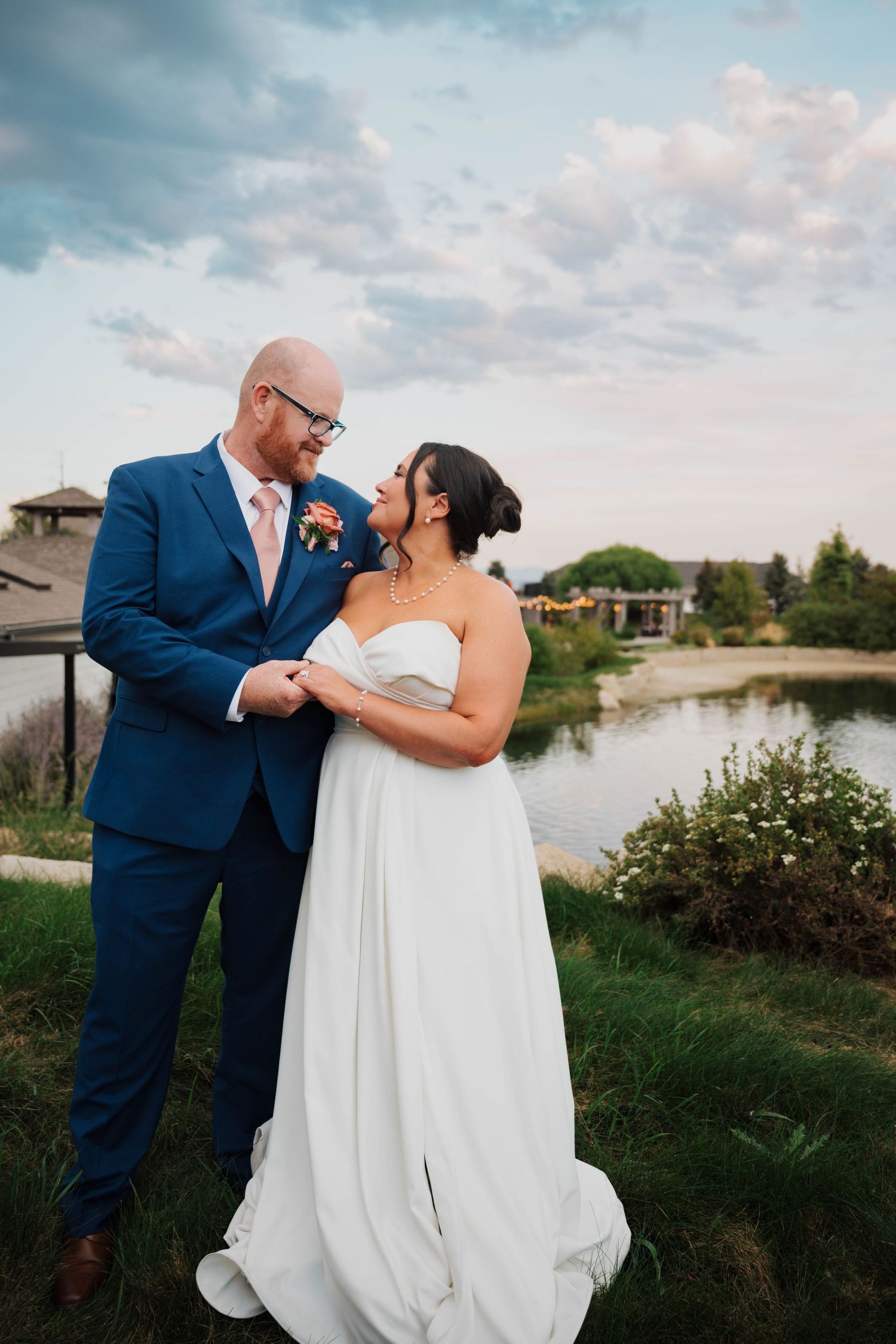 Bride in white strapless gown and groom in blue suit share a romantic moment outdoors near a pond at golden hour.