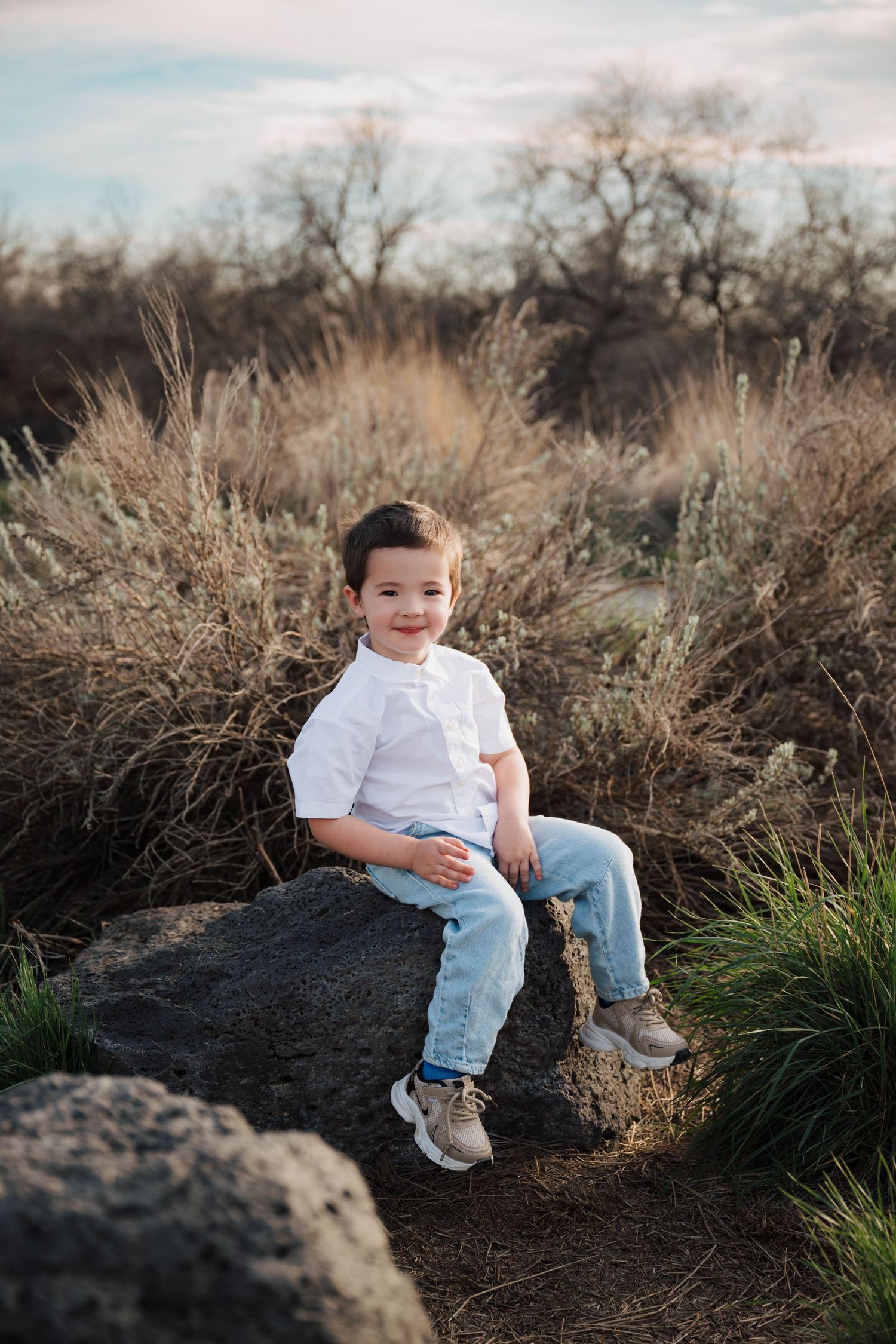 A smiling young boy in a white shirt and jeans sits on a rock surrounded by dry grass and shrubs outdoors.