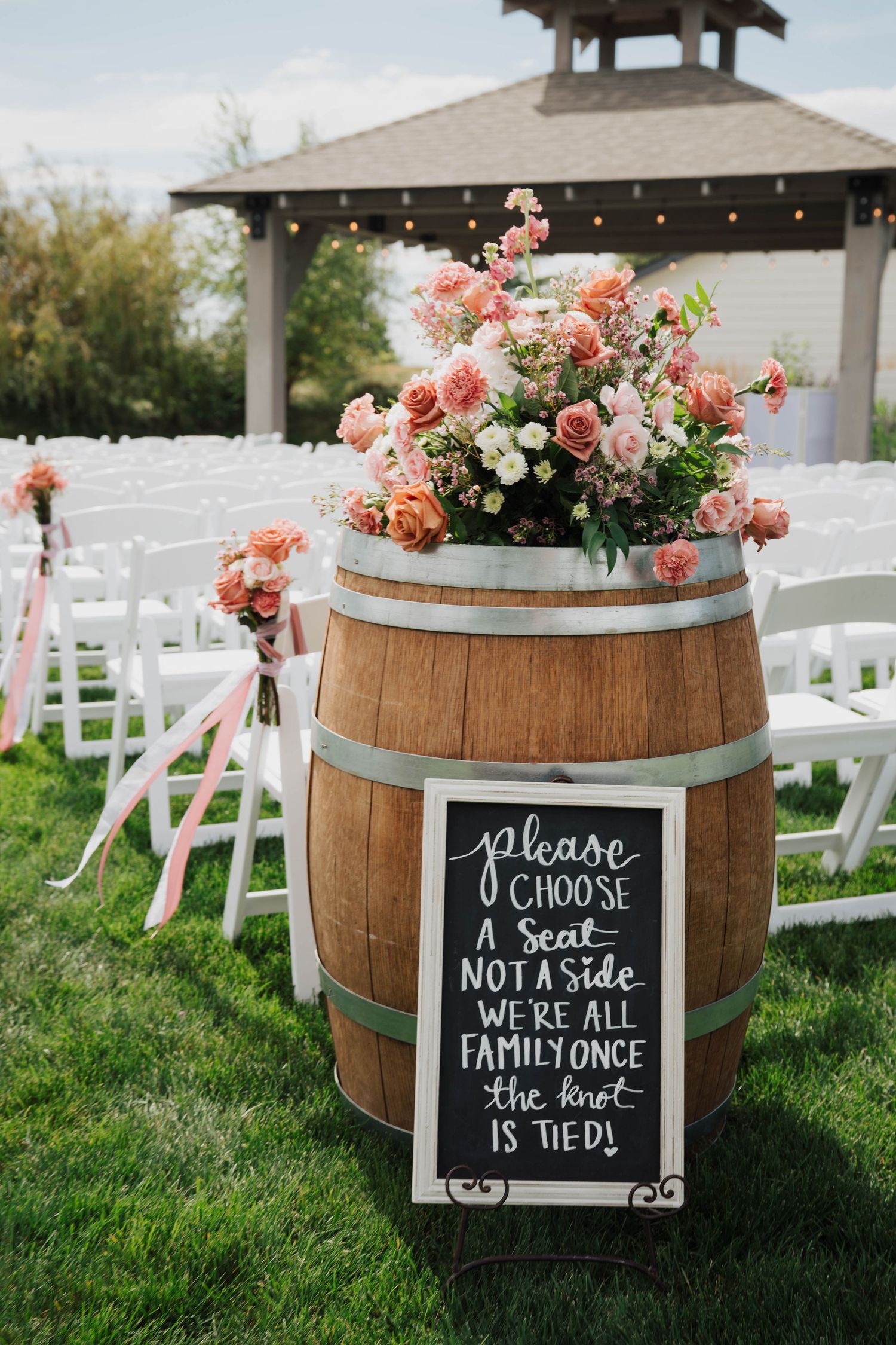 Rustic wedding ceremony setup with a wooden barrel topped with pink roses and a chalkboard sign reading please choose a seat not a side.