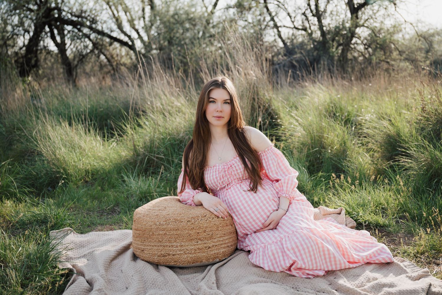 Pregnant woman in pink dress sitting on blanket in nature.