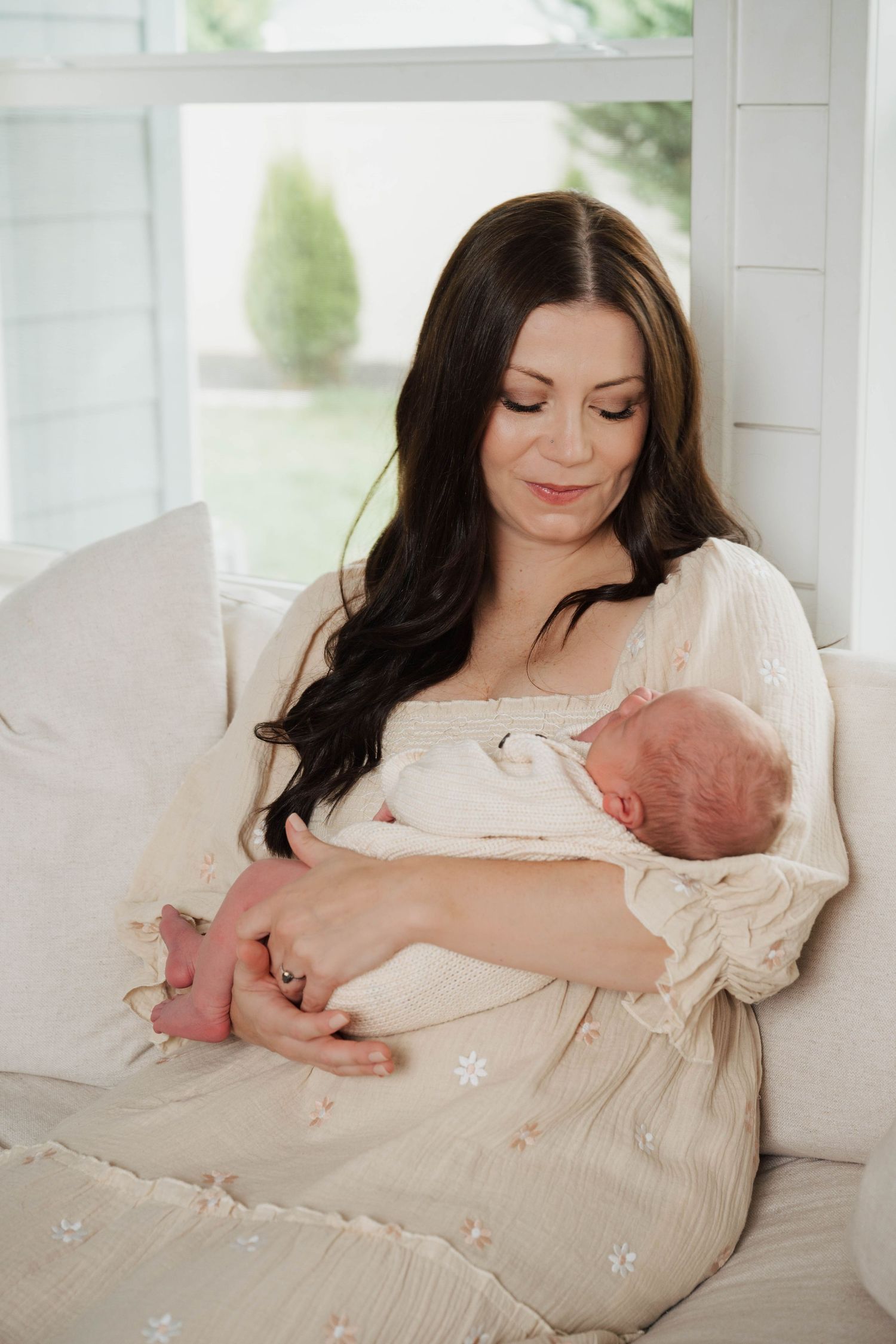 Mother in white dress holding newborn baby on cream couch near window.