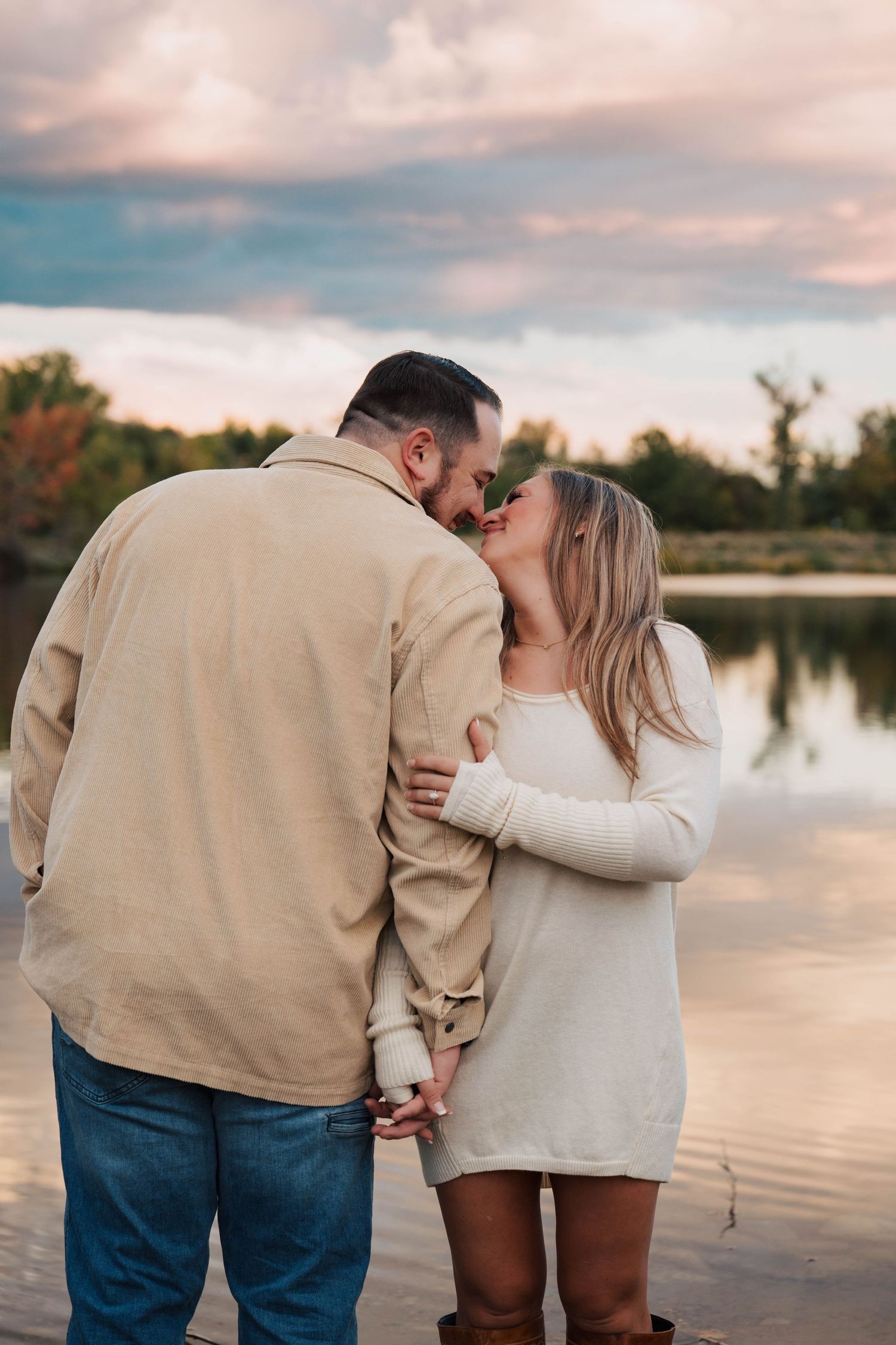 A romantic couple embraces near a serene lake during golden hour.