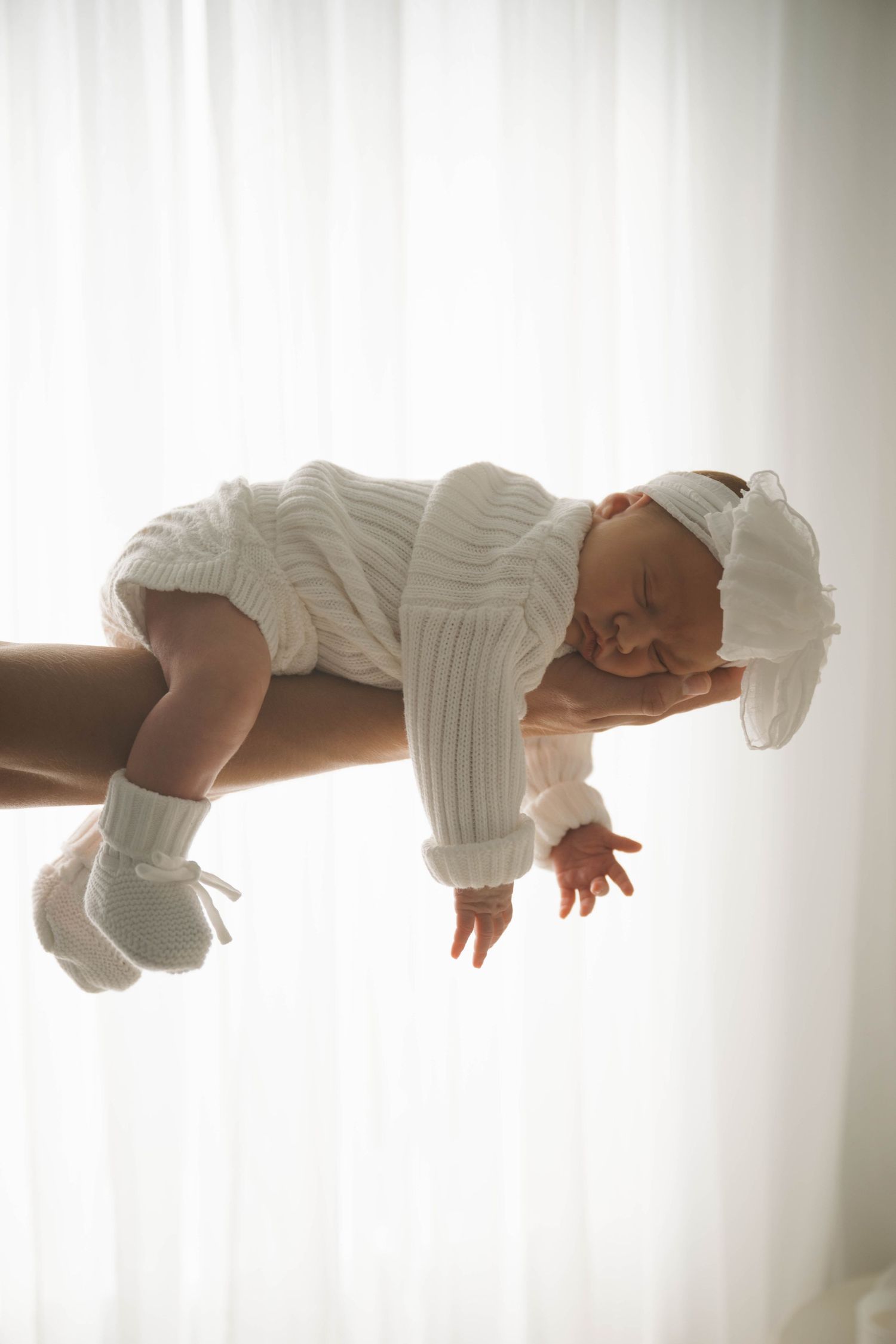 Newborn baby in white knit outfit and headband resting on adult hand against soft white curtains.