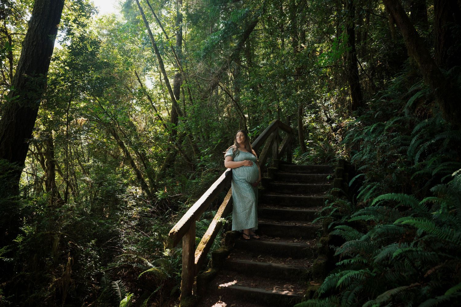 Pregnant woman in a sage green dress standing on wooden forest stairs surrounded by lush green trees and ferns.