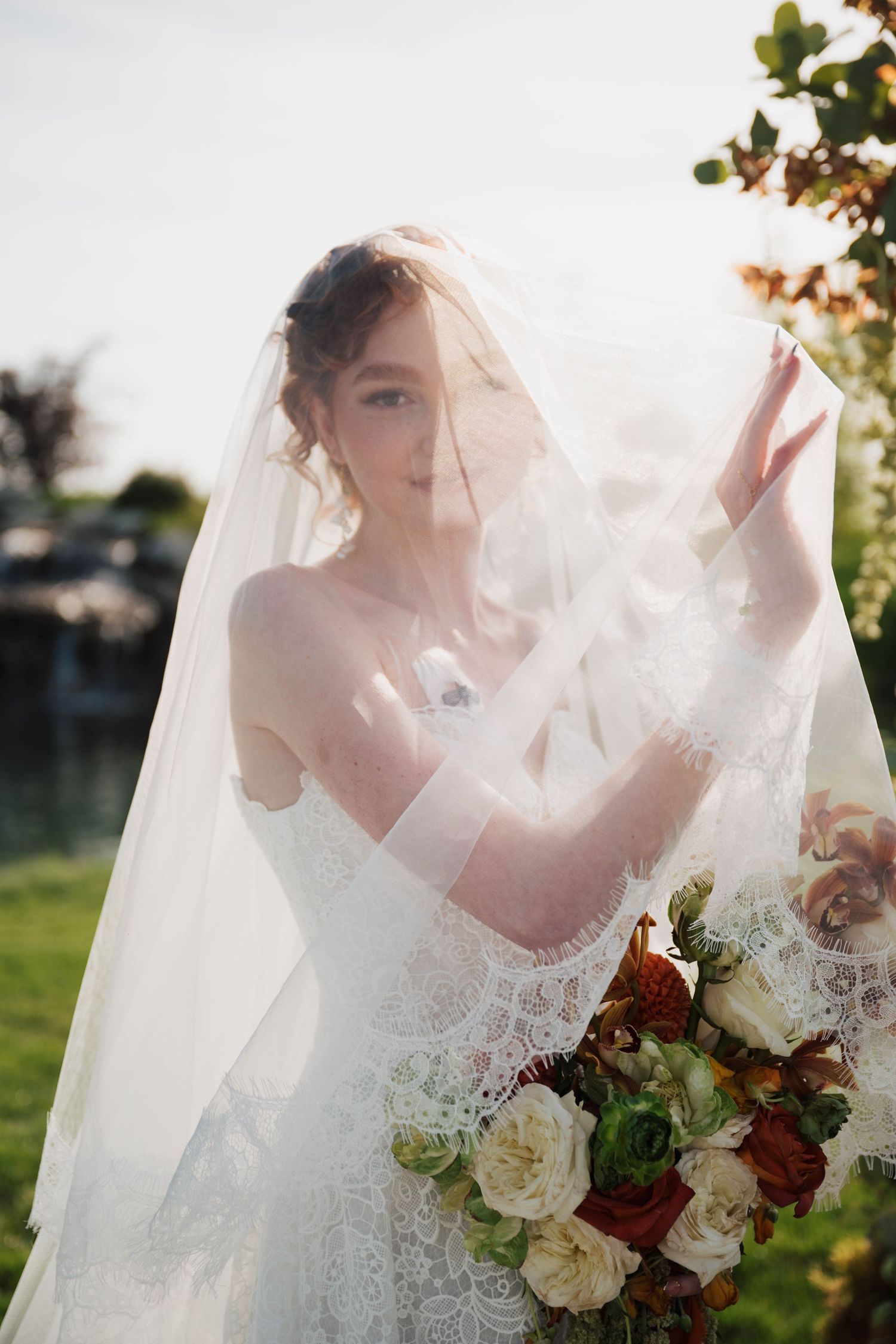 Bride in lace wedding dress and veil holding a bouquet of red and white roses outdoors in natural light.