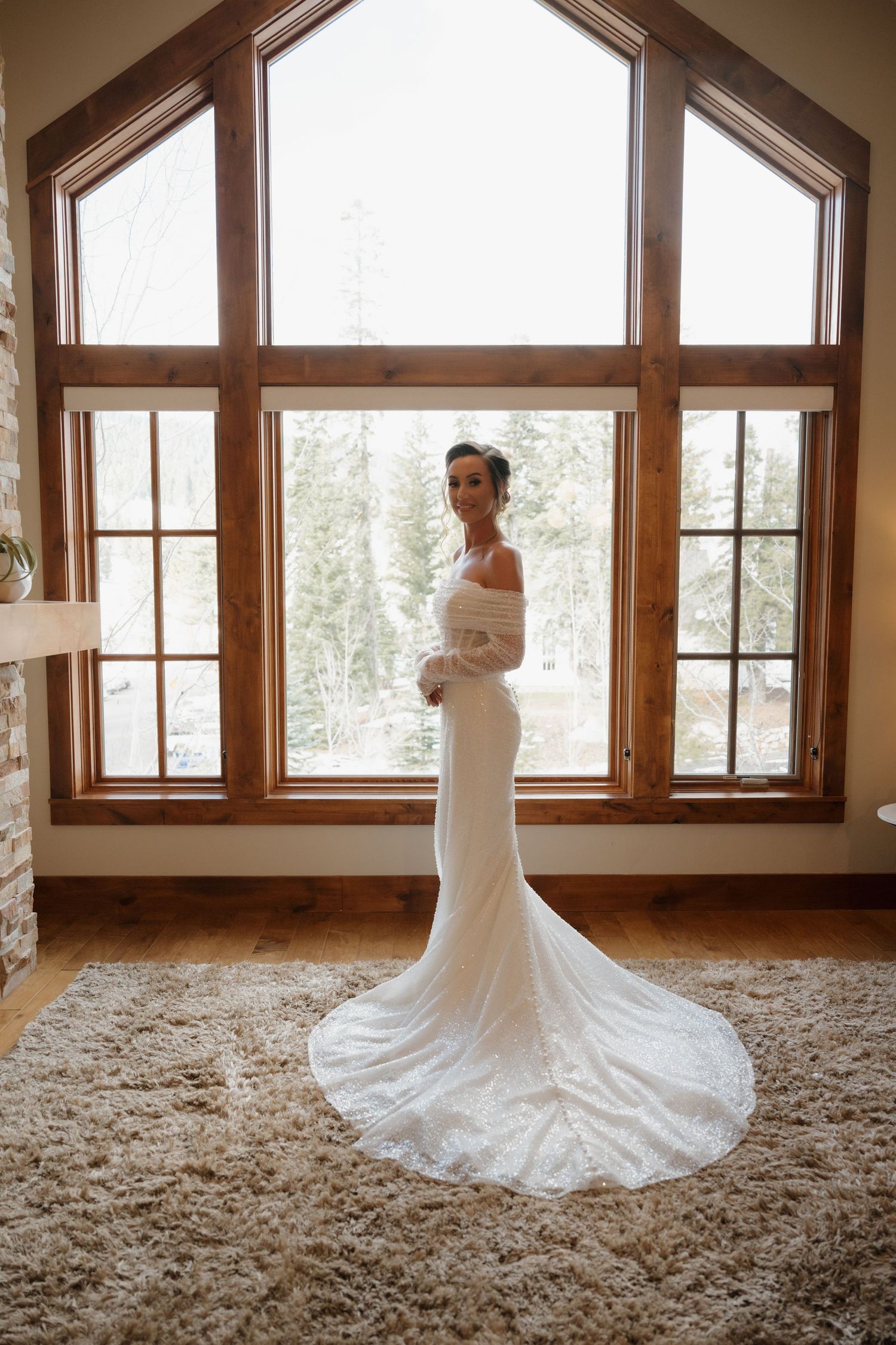 A white wedding dress with a long train displayed in a rustic room with large windows and wooden beams.