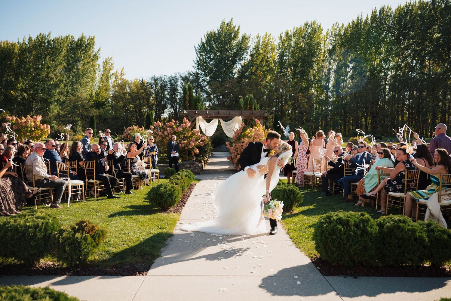 Bride and groom kiss at outdoor wedding ceremony aisle surrounded by guests and lush green trees at the chateau des fleurs in eagle, idaho