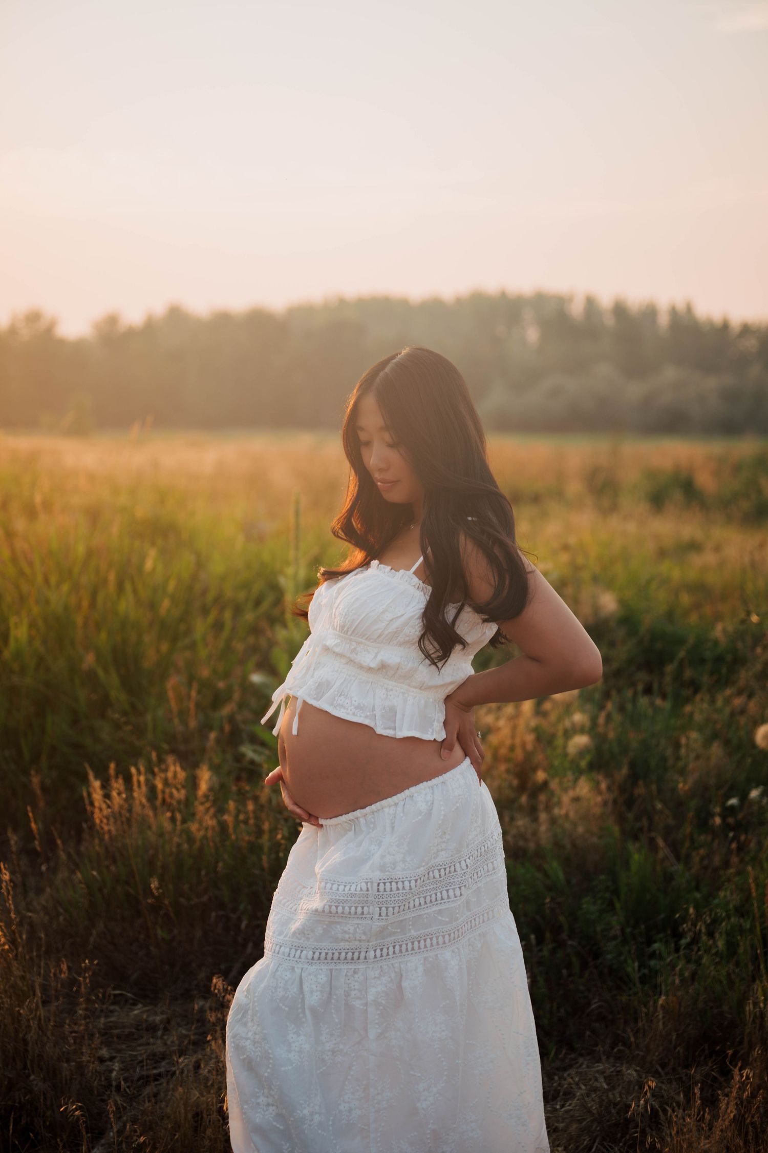 Pregnant woman in white crop top and skirt holding her baby bump in a golden sunset field.