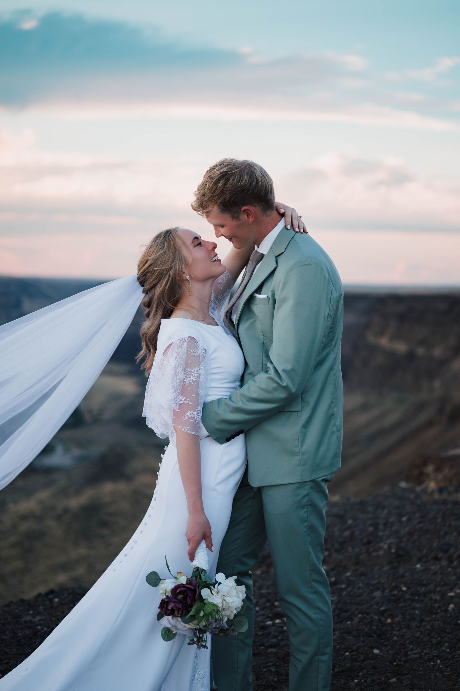 Bride and groom share a romantic kiss outdoors at sunset, with veil flowing in the wind and scenic landscape backdrop.