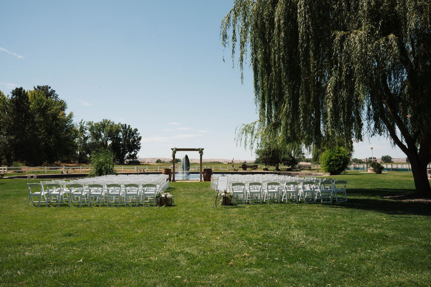 A panoramic view of a green lawn wedding ceremony setup with white chairs under weeping willow trees.