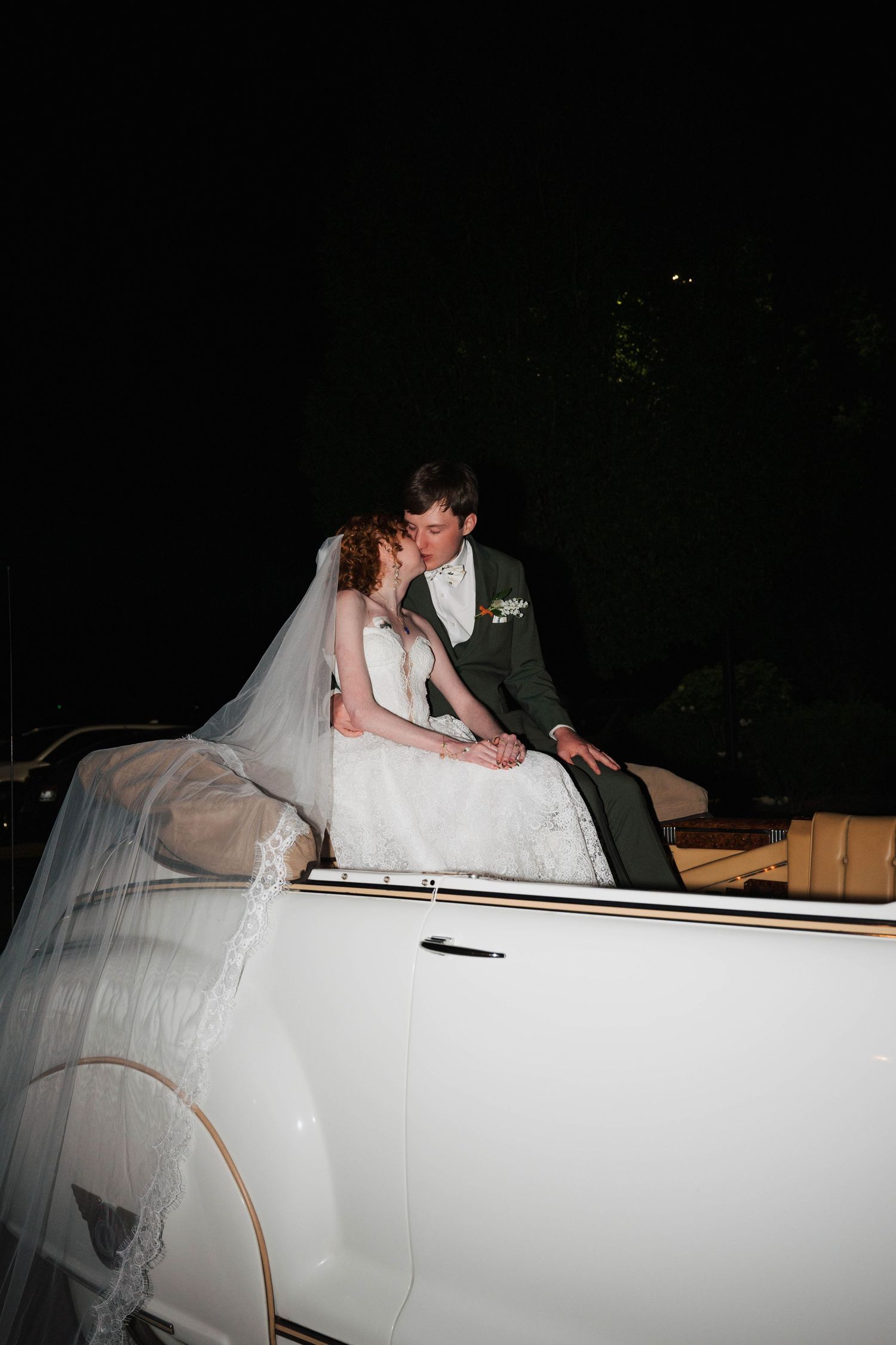 Bride and groom share romantic kiss seated on classic white convertible car during nighttime wedding celebration.