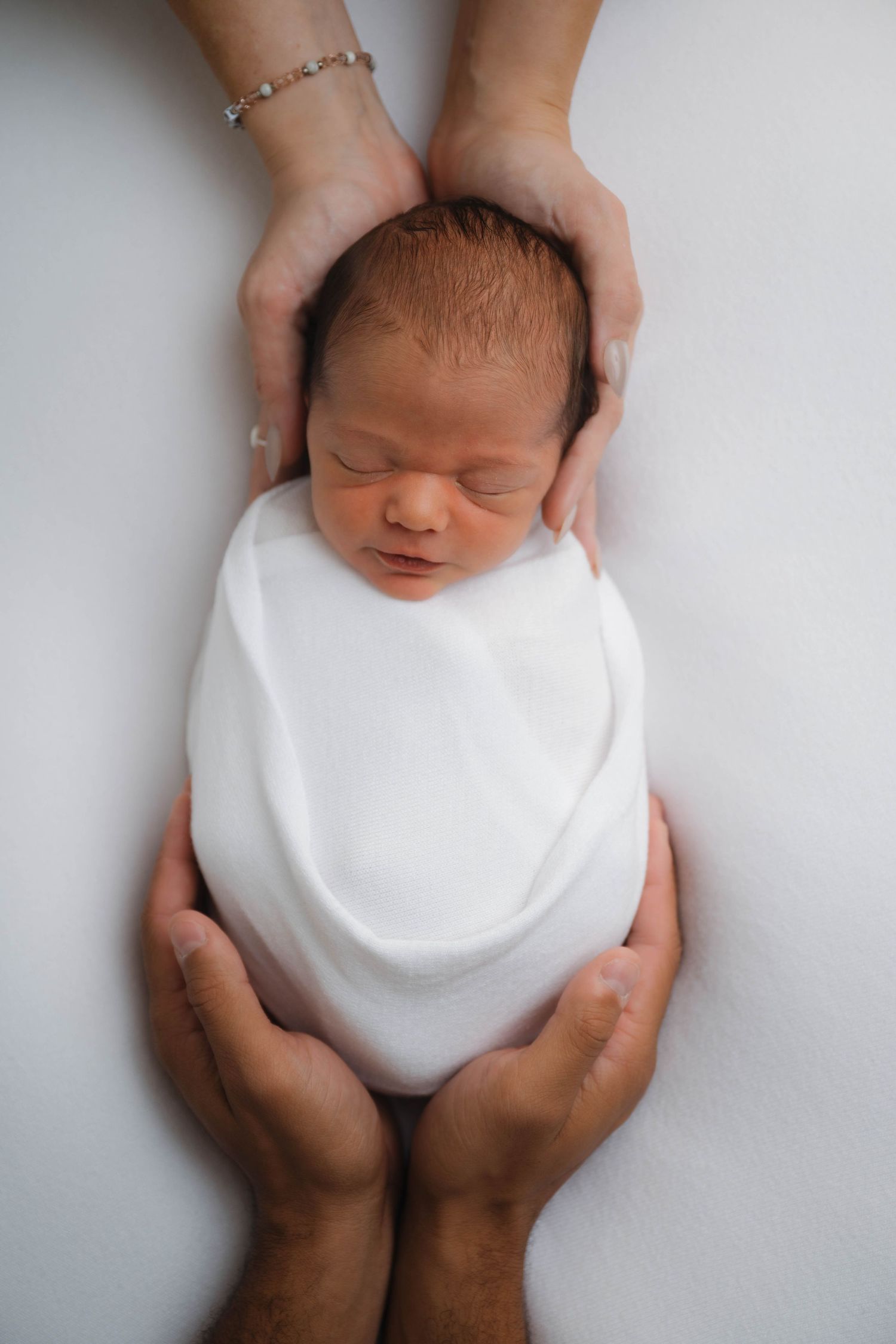 Newborn baby swaddled in white wrap, cradled gently by two pairs of adult hands on a soft white background.
