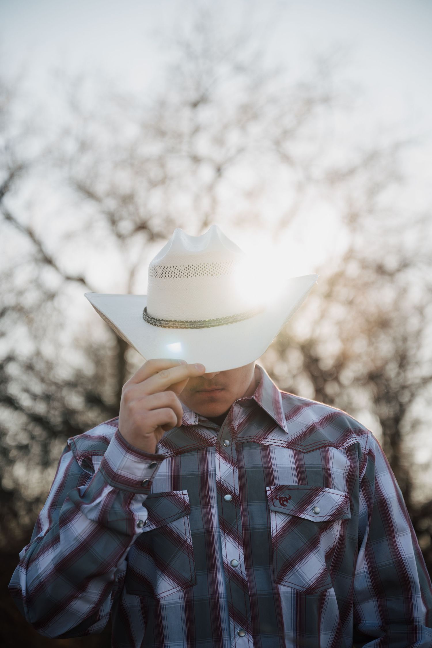 A person in a plaid western shirt and cowboy hat stands against a sunset backdrop.
