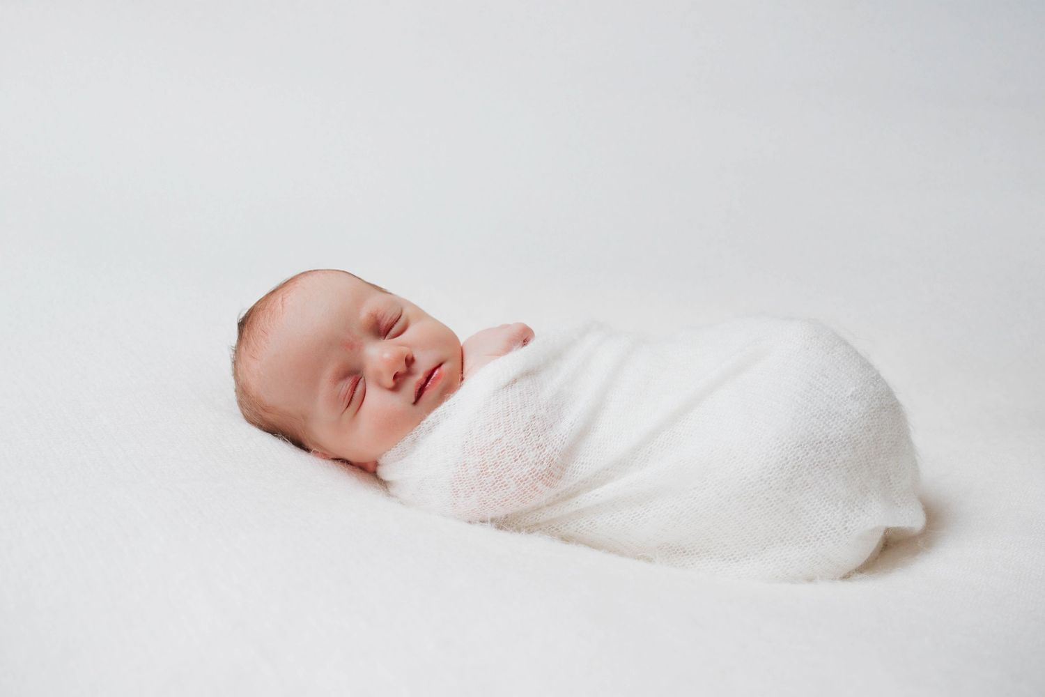 Sleeping newborn baby wrapped in soft white blanket on light background.