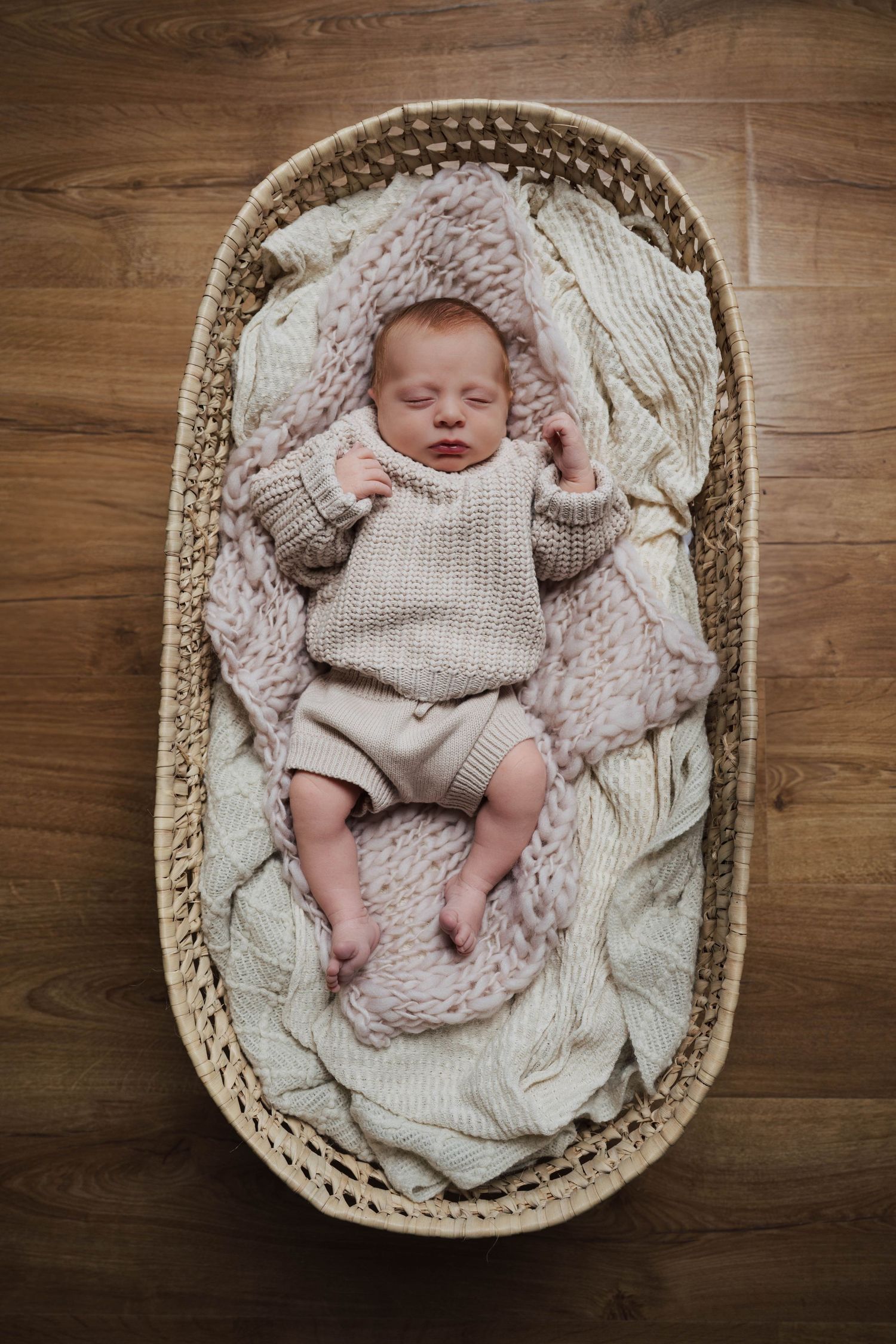 Sleeping newborn baby nestled in cozy woven basket on wooden floor with soft knit blankets.