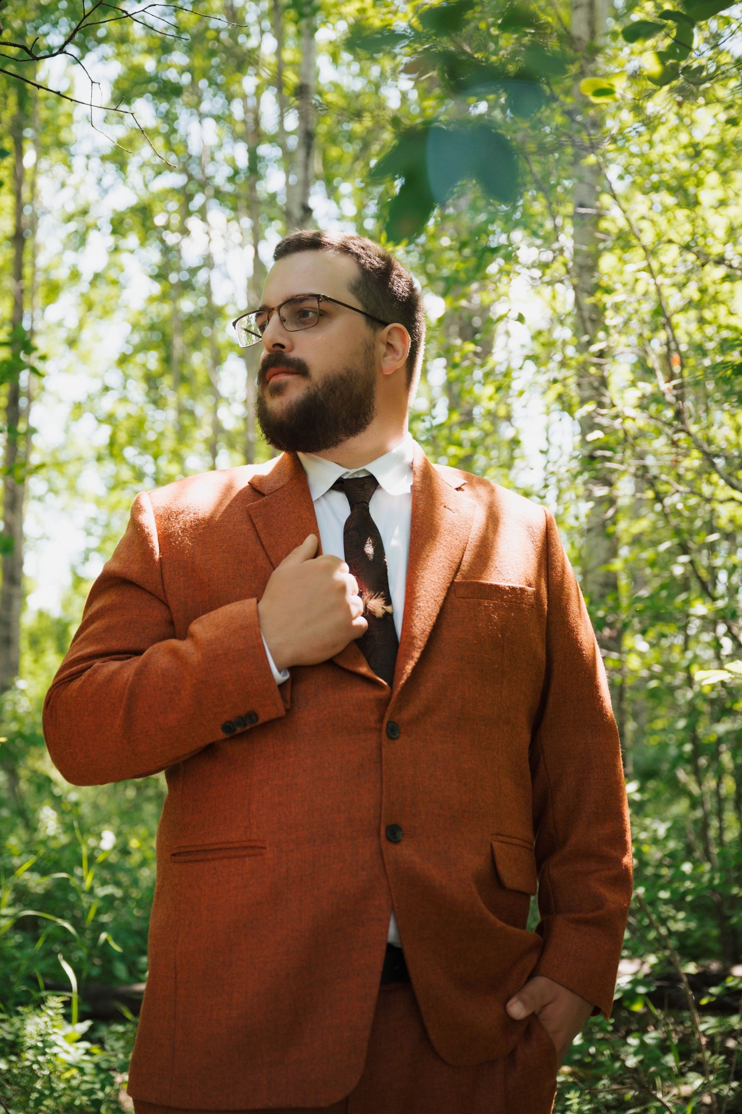 A groom in a stylish rust-colored suit poses in a sunlit forest setting.