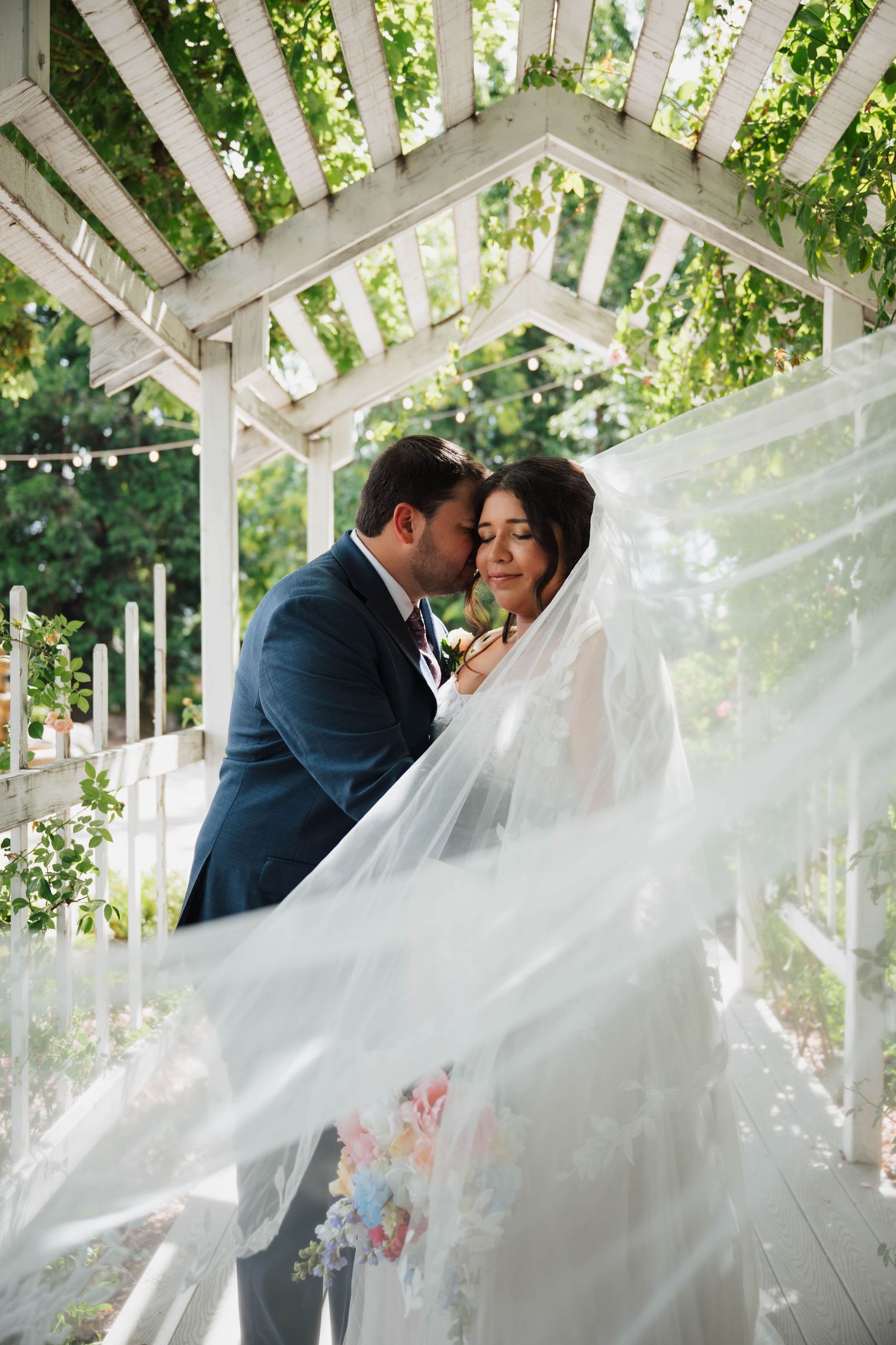 Bride and groom embrace under white pergola with flowing veil, surrounded by lush greenery at outdoor wedding ceremony.