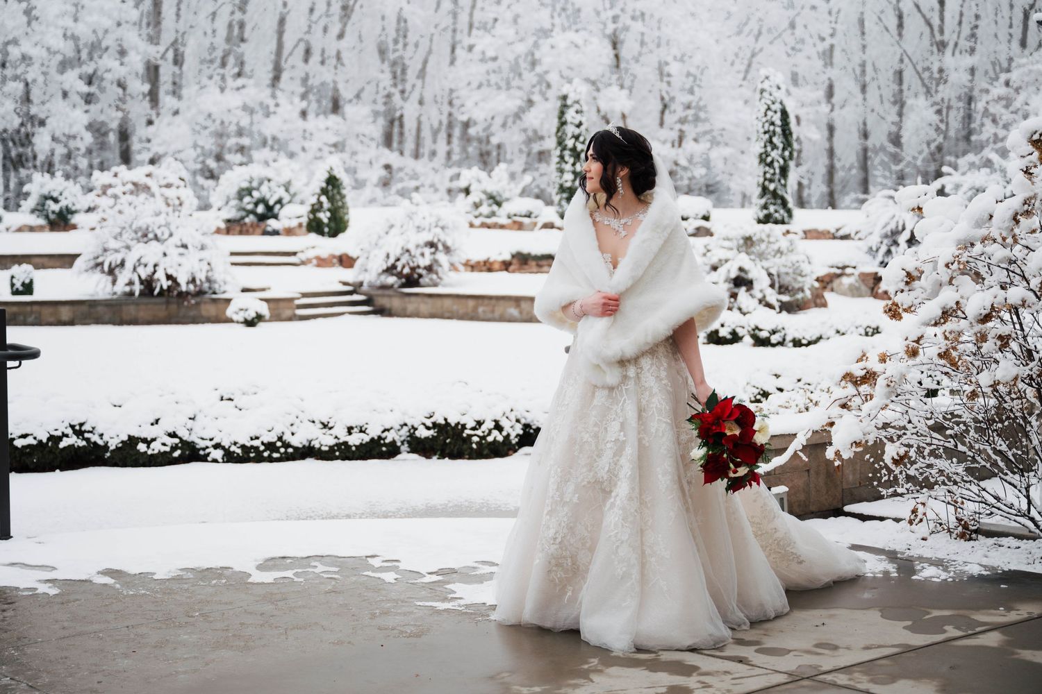 Bride in white lace gown and fur wrap holding red bouquet in snowy winter garden wedding setting.