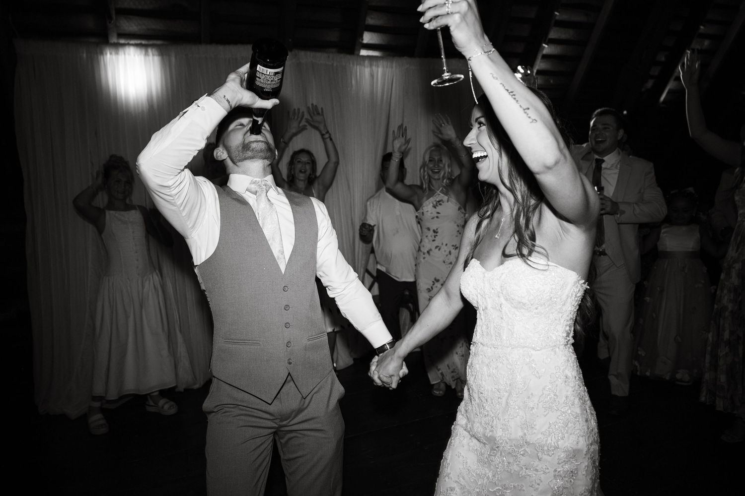 Black and white photo of bride and groom dancing and raising glasses at their wedding reception.