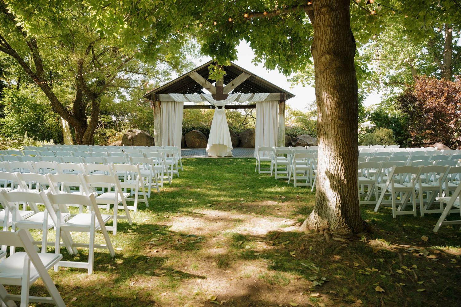 White wooden gazebo decorated with flowing curtains set up for an outdoor wedding ceremony with rows of folding chairs.