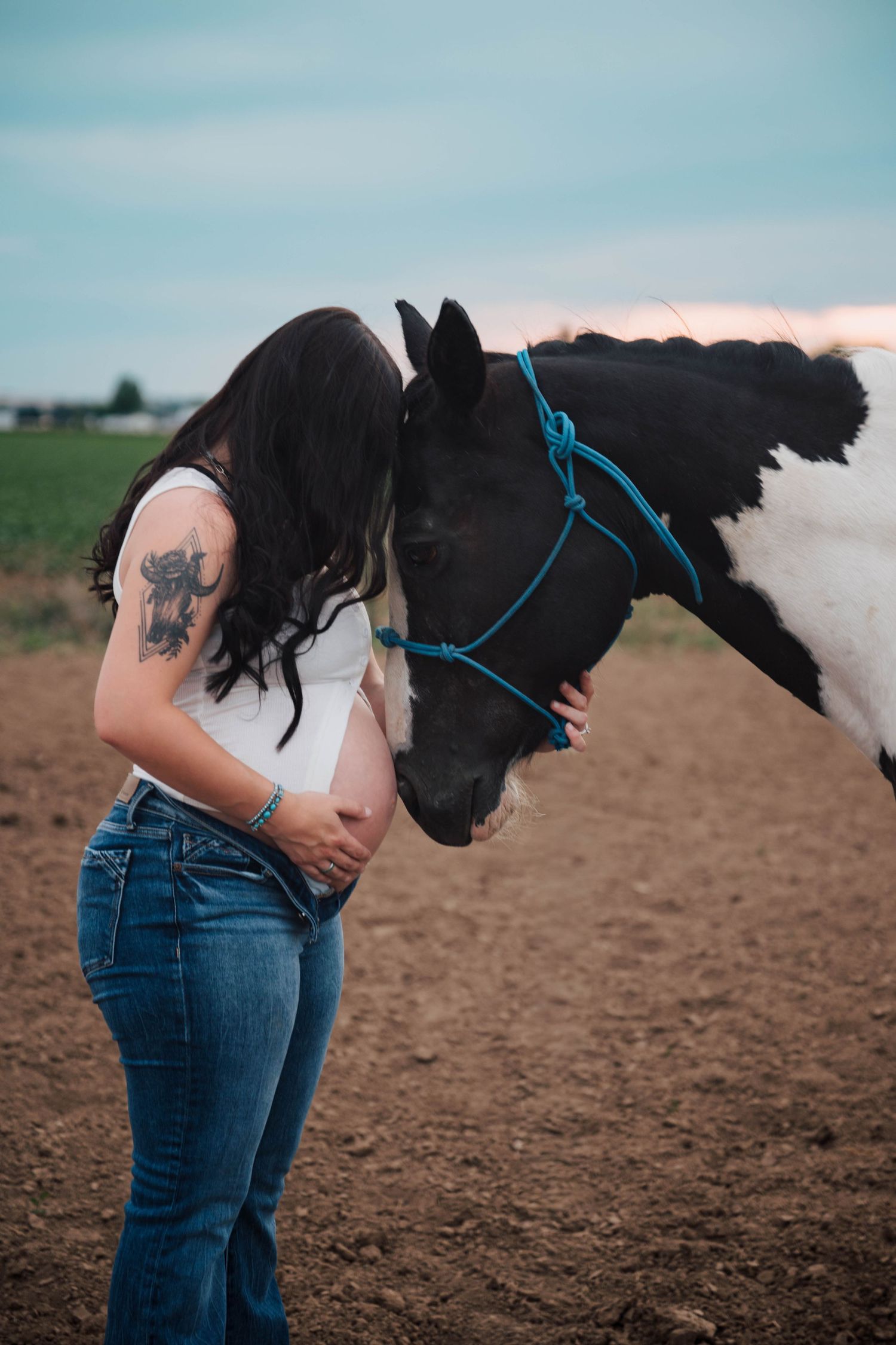 Pregnant woman with tattoos nuzzles a black and white horse wearing a blue halter in an open field at dusk.