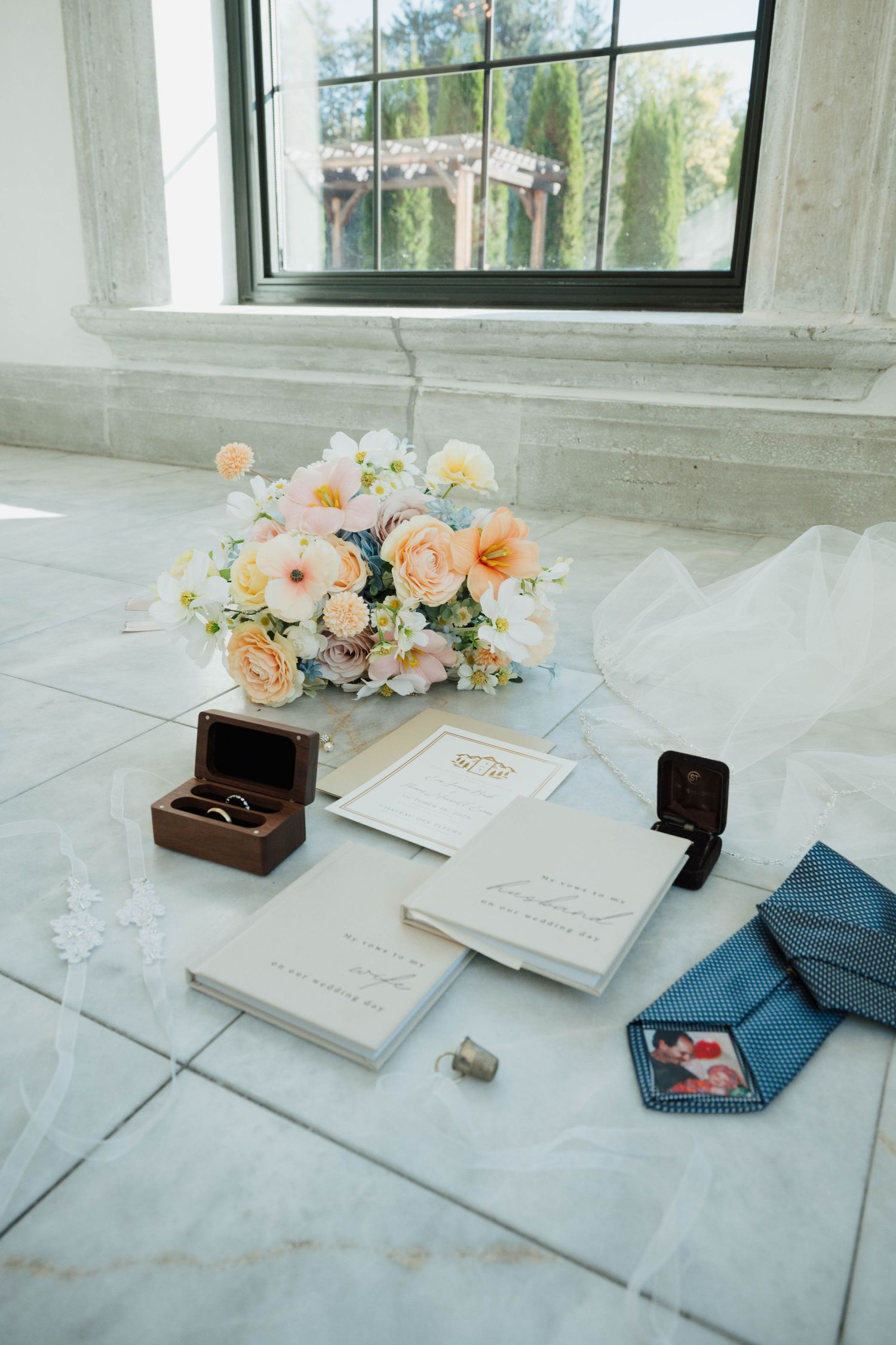 Wedding flat lay with peach floral bouquet, invitation suite, ring box, blue tie, and vow books on marble floor.