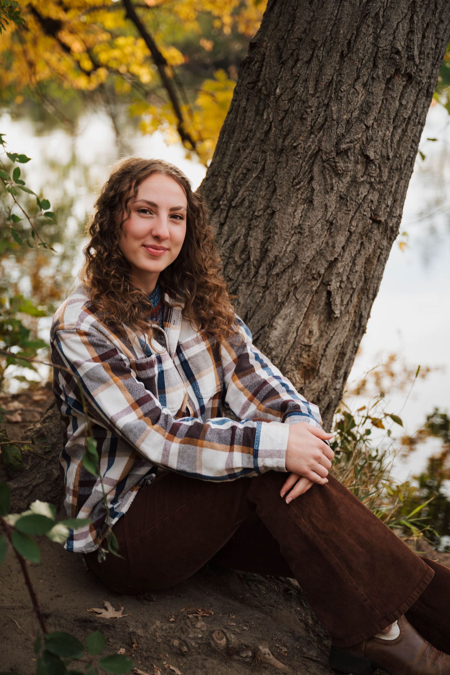 Person in plaid shirt relaxing against a large tree trunk with fall colors in background.