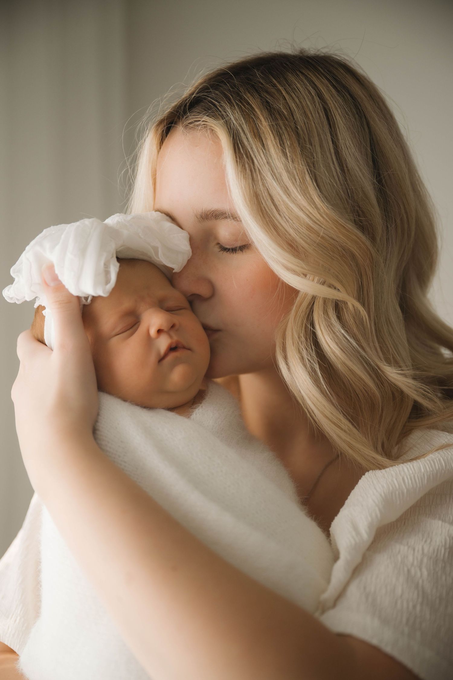 Blonde mother tenderly kissing her sleeping newborn baby wrapped in white cloth with a bow headband.