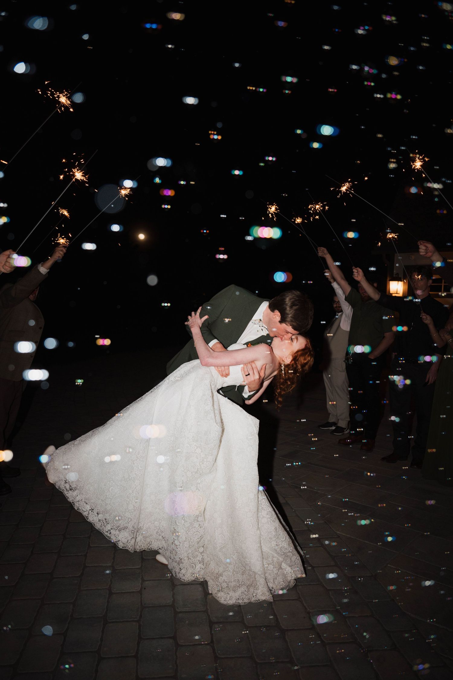 Bride and groom share a romantic dip kiss surrounded by sparklers and bokeh lights at a nighttime wedding sendoff.