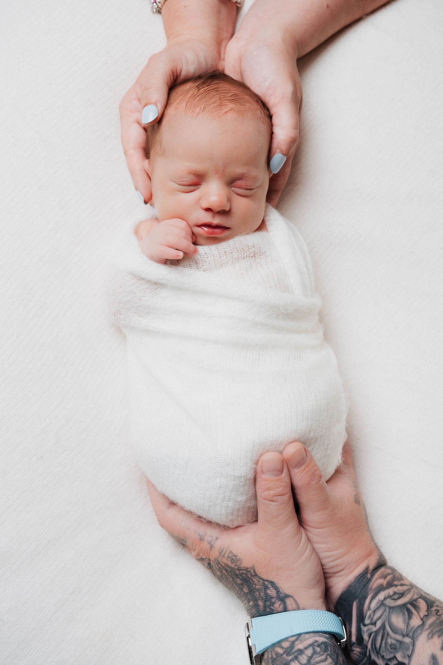 Newborn baby wrapped in white blanket with tattooed hands gently holding.