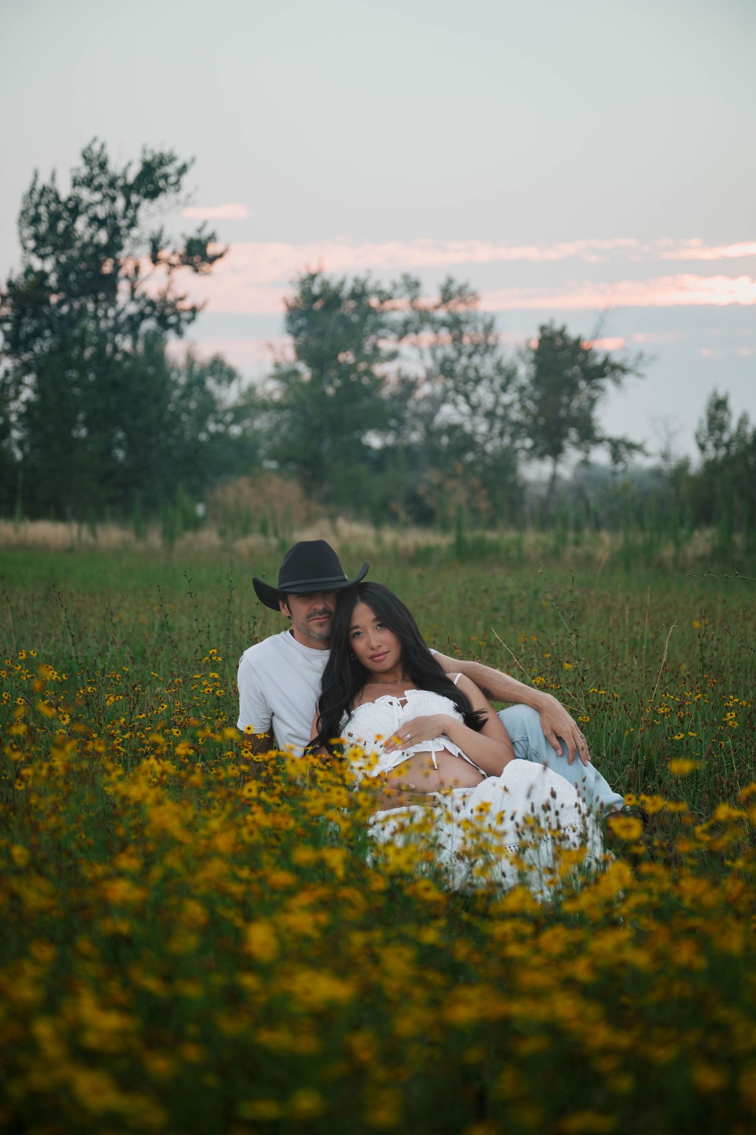 Couple sitting in yellow wildflower field at sunset, man in cowboy hat embracing woman in white dress.
