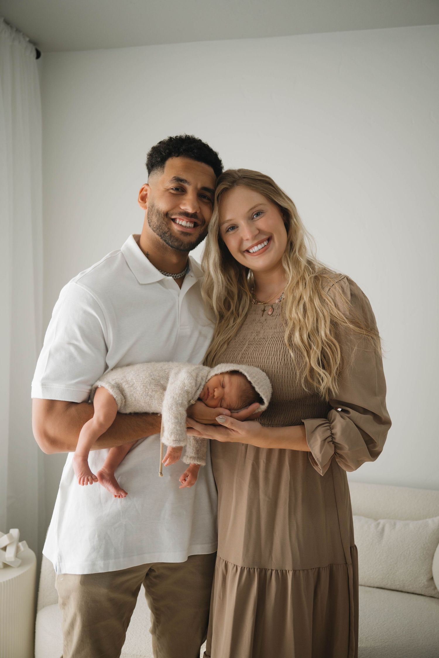 Happy couple smiling and holding their newborn baby in a bright, cozy room during a professional newborn photo session.
