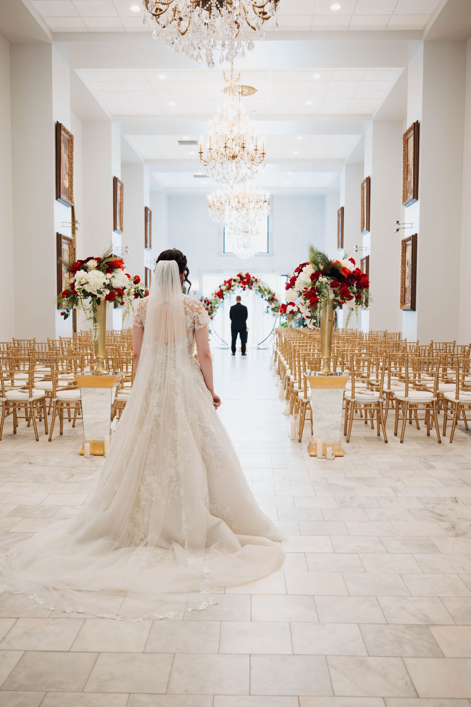 Bride in elegant lace gown with long veil walks down aisle of chandelier-lit venue with red floral arch.