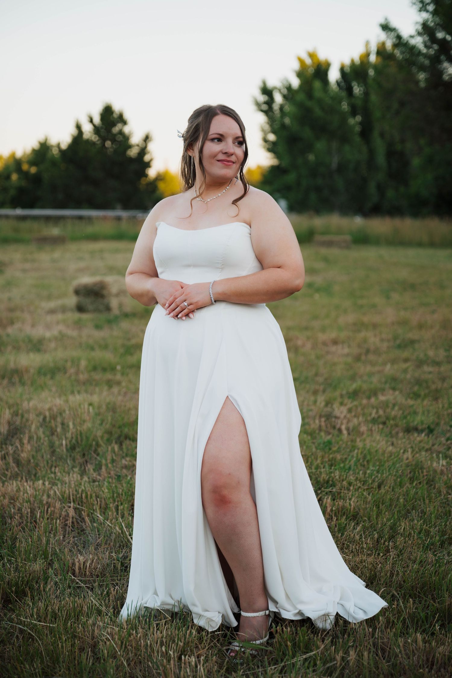 A plus-size bride in a white strapless gown with a thigh-high slit poses in a golden field at sunset.