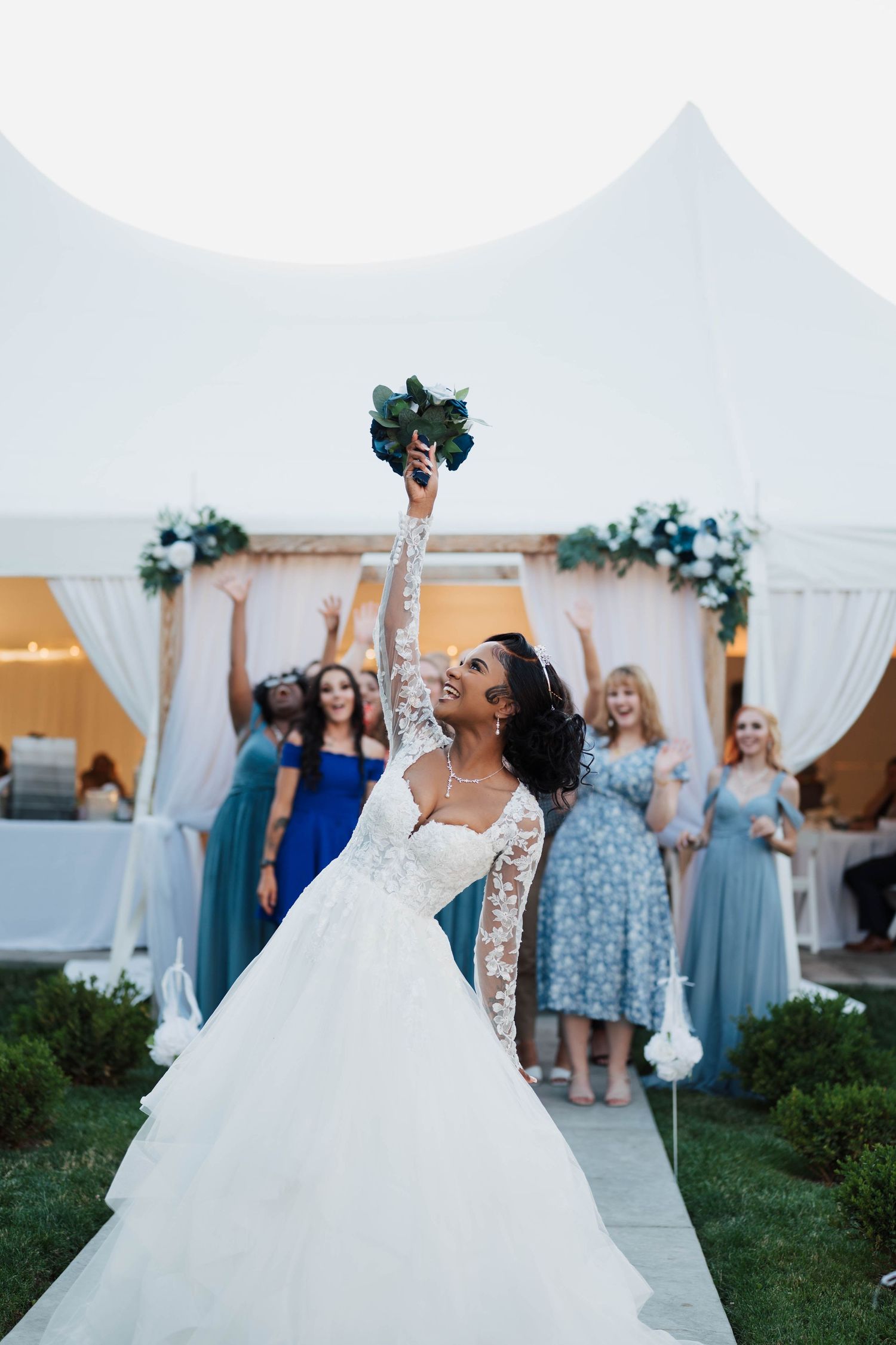 Bride in white lace gown tossing bouquet at outdoor wedding reception with bridesmaids in blue dresses cheering behind her.