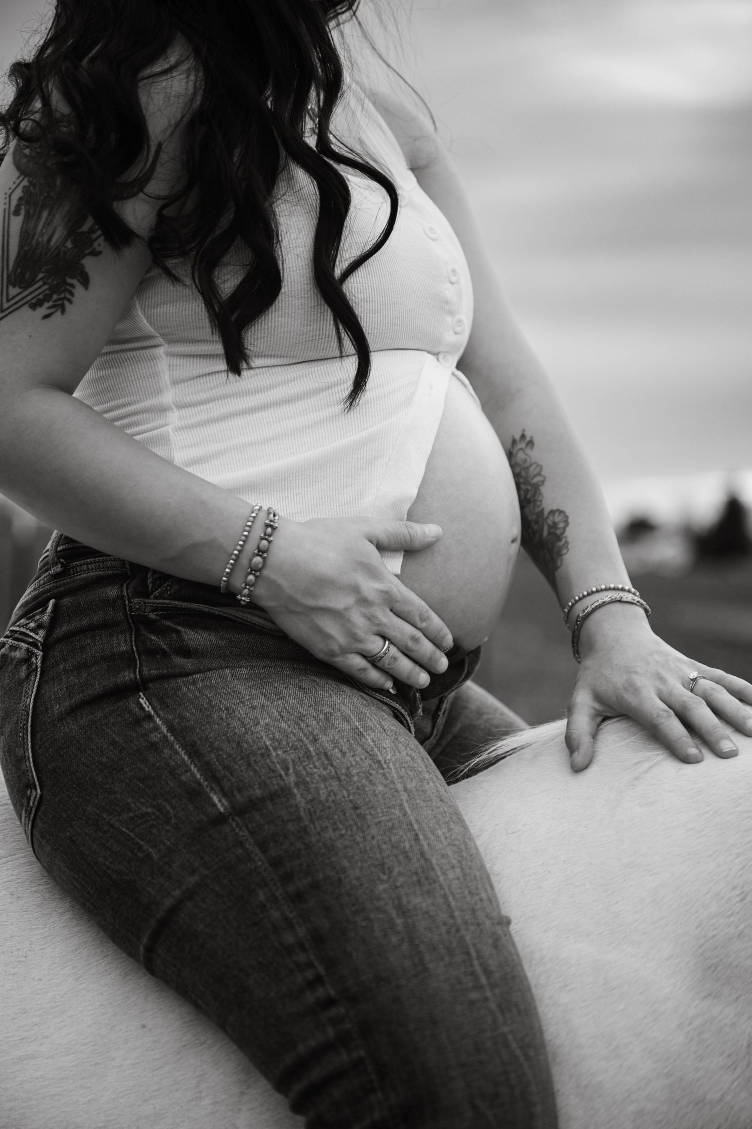 Black and white maternity photo of pregnant woman in jeans cradling baby bump, with tattoos and bracelets visible.