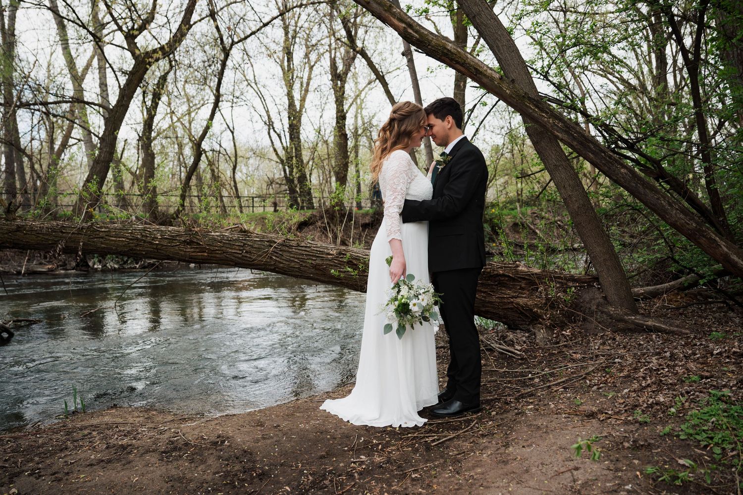 A couple in wedding attire stands near a wooded stream during their outdoor photoshoot.
