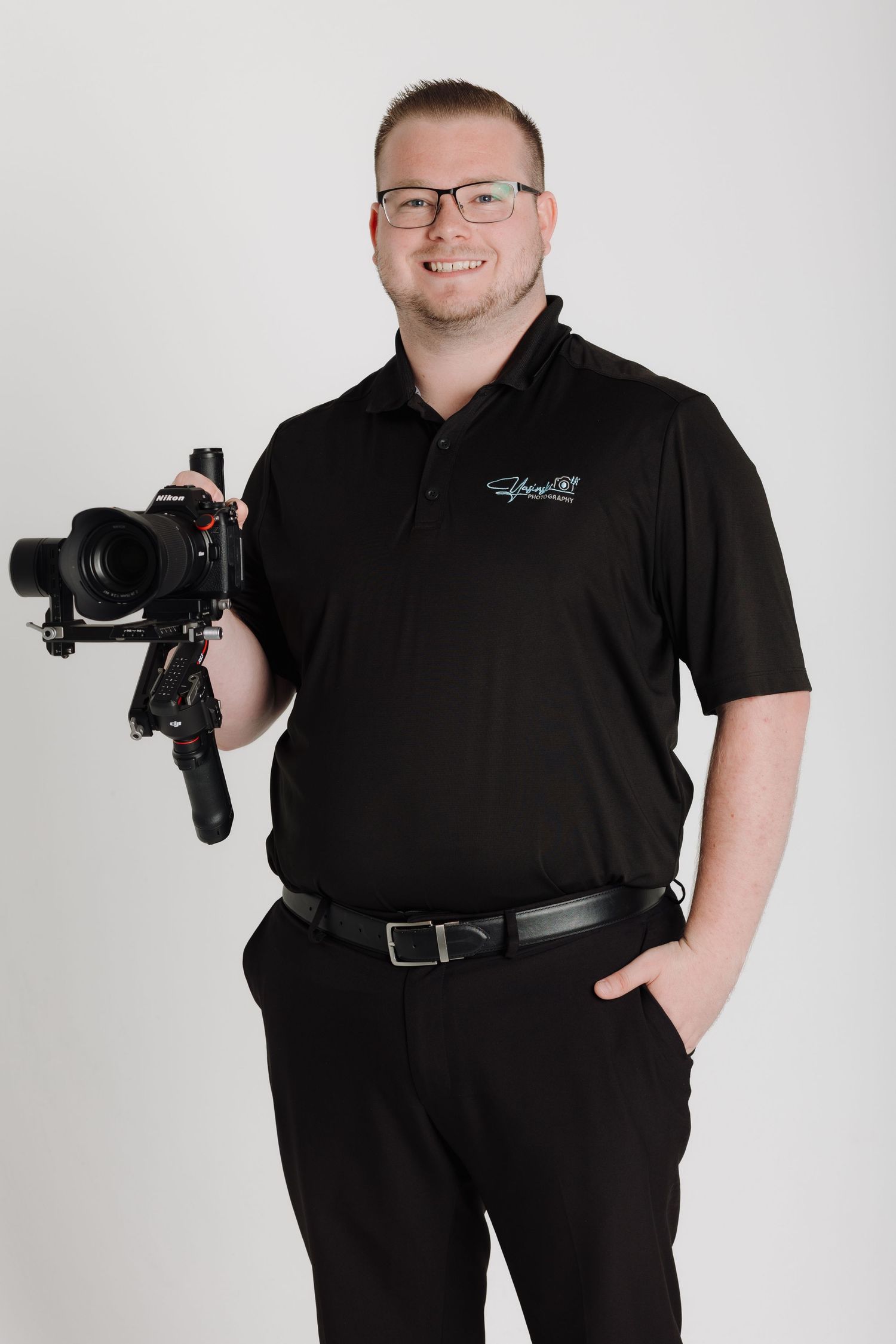 Professional photographer in black attire posing with camera gear against white backdrop.