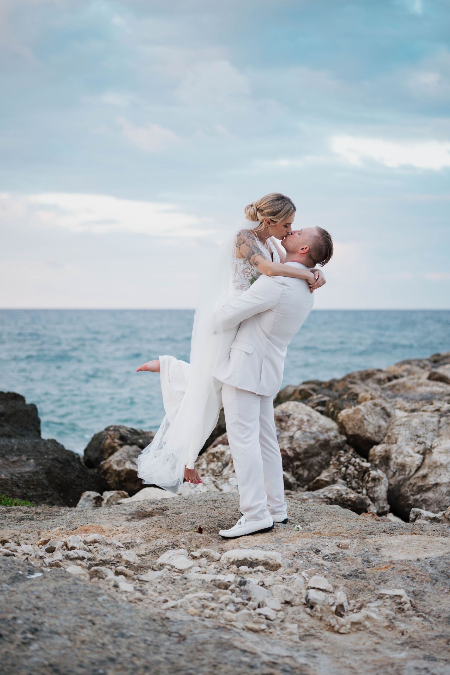 Groom in white suit lifts bride in lace wedding dress for a kiss on rocky ocean shore at sunset.