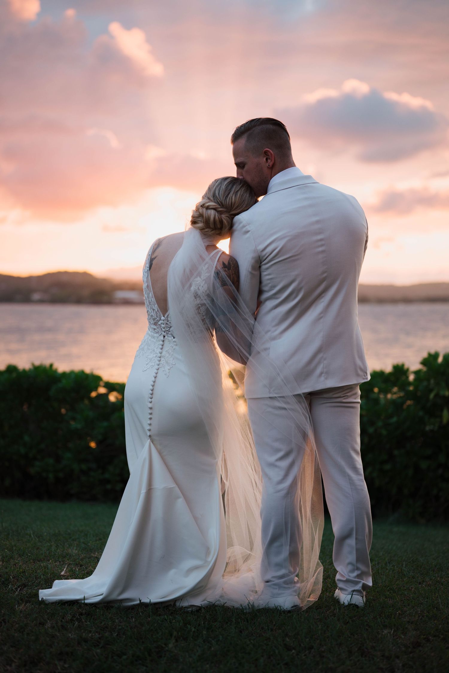 Bride and groom embrace at sunset by the ocean, wearing white wedding attire with a pink sky backdrop.