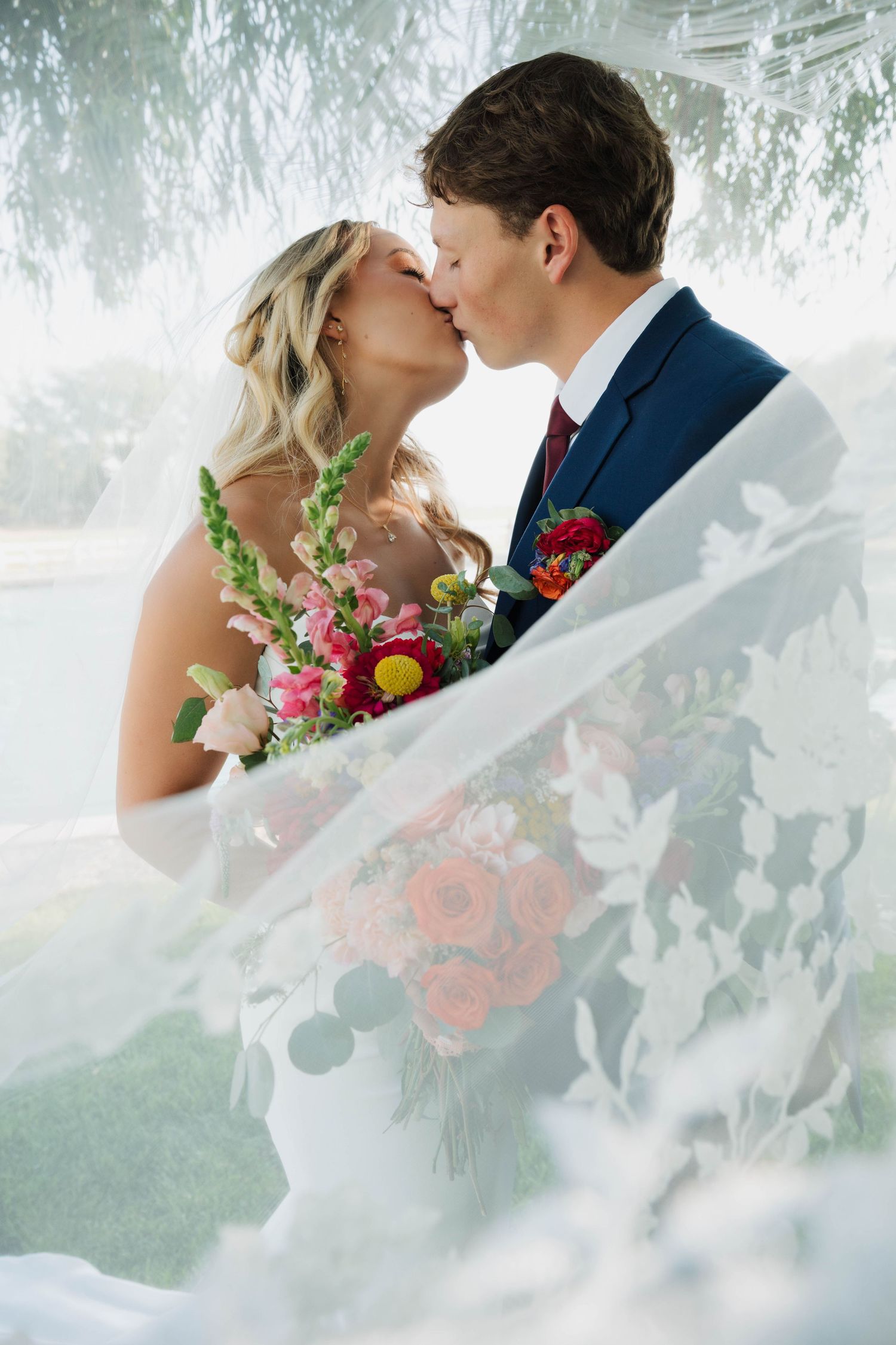 Bride and groom share a romantic kiss under a willow tree, bride holding a vibrant bouquet, veil flowing in foreground.