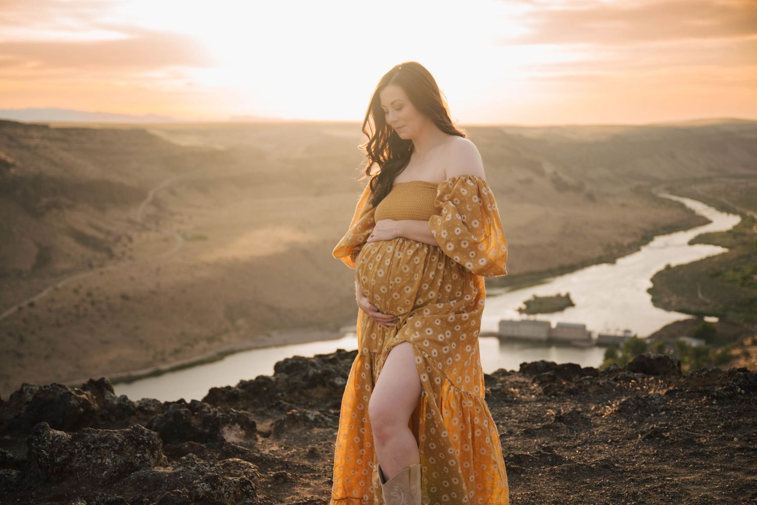 Woman in flowing yellow dress poses against dramatic sunset landscape with river view.