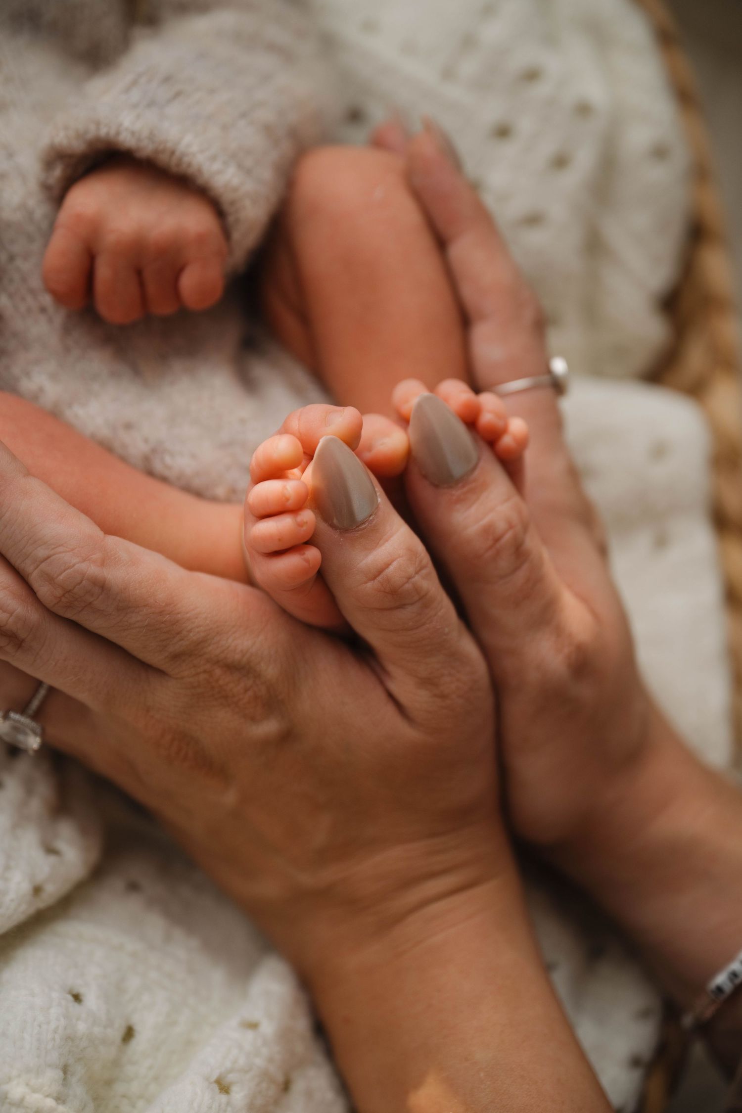 Close-up of adult hands with taupe nails gently holding tiny newborn baby feet wrapped in a white knit blanket.