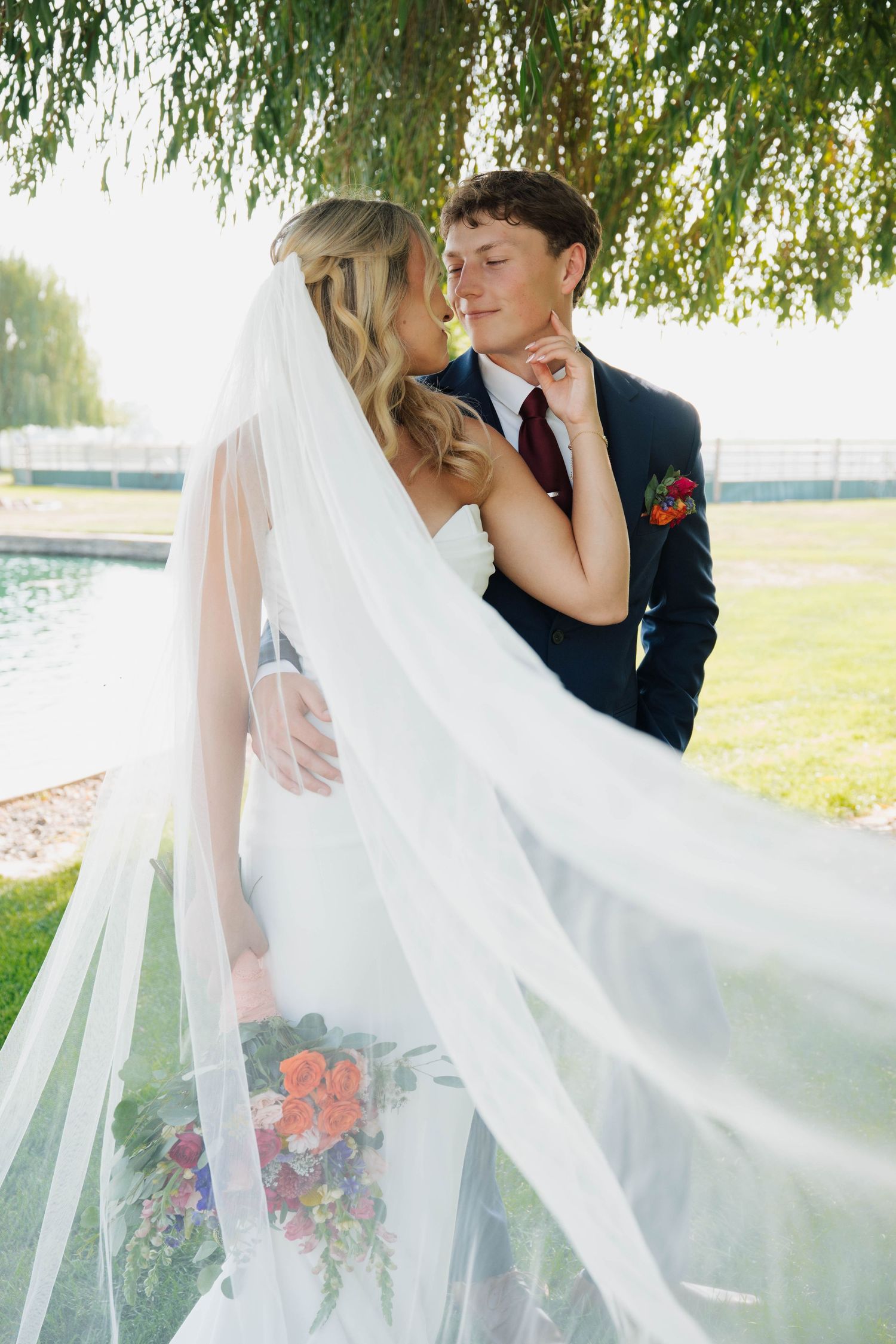 Bride and groom embrace under a weeping willow tree, veil flowing, holding a colorful bouquet.