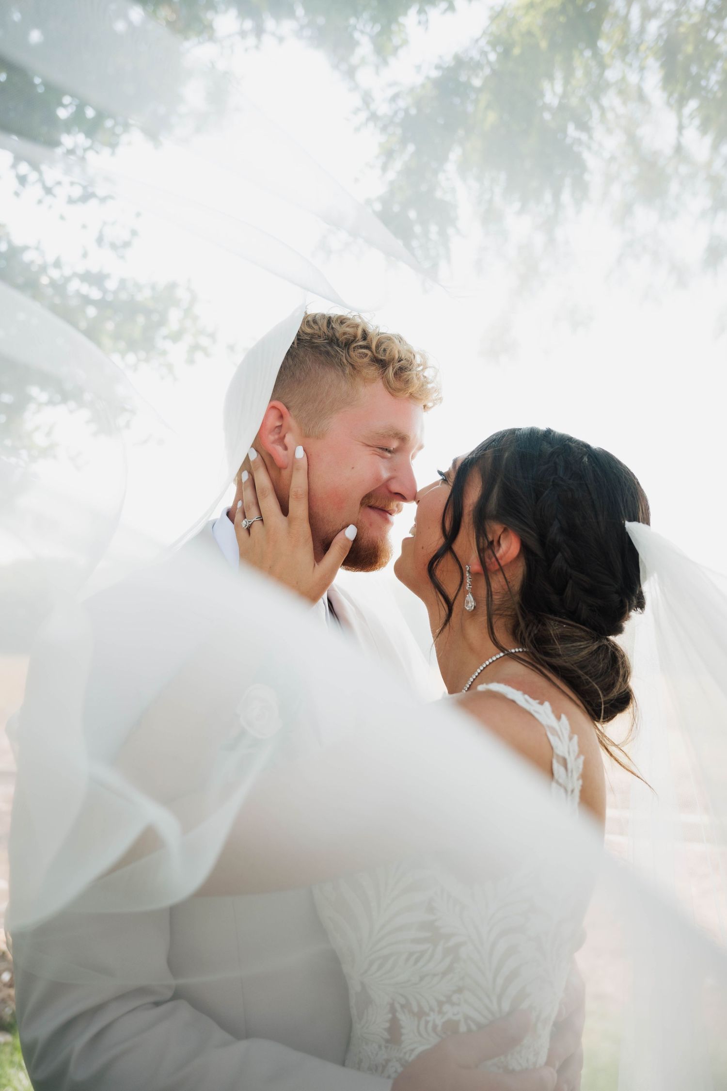 A joyful bride and groom share an intimate moment under a flowing veil, smiling and touching foreheads outdoors.