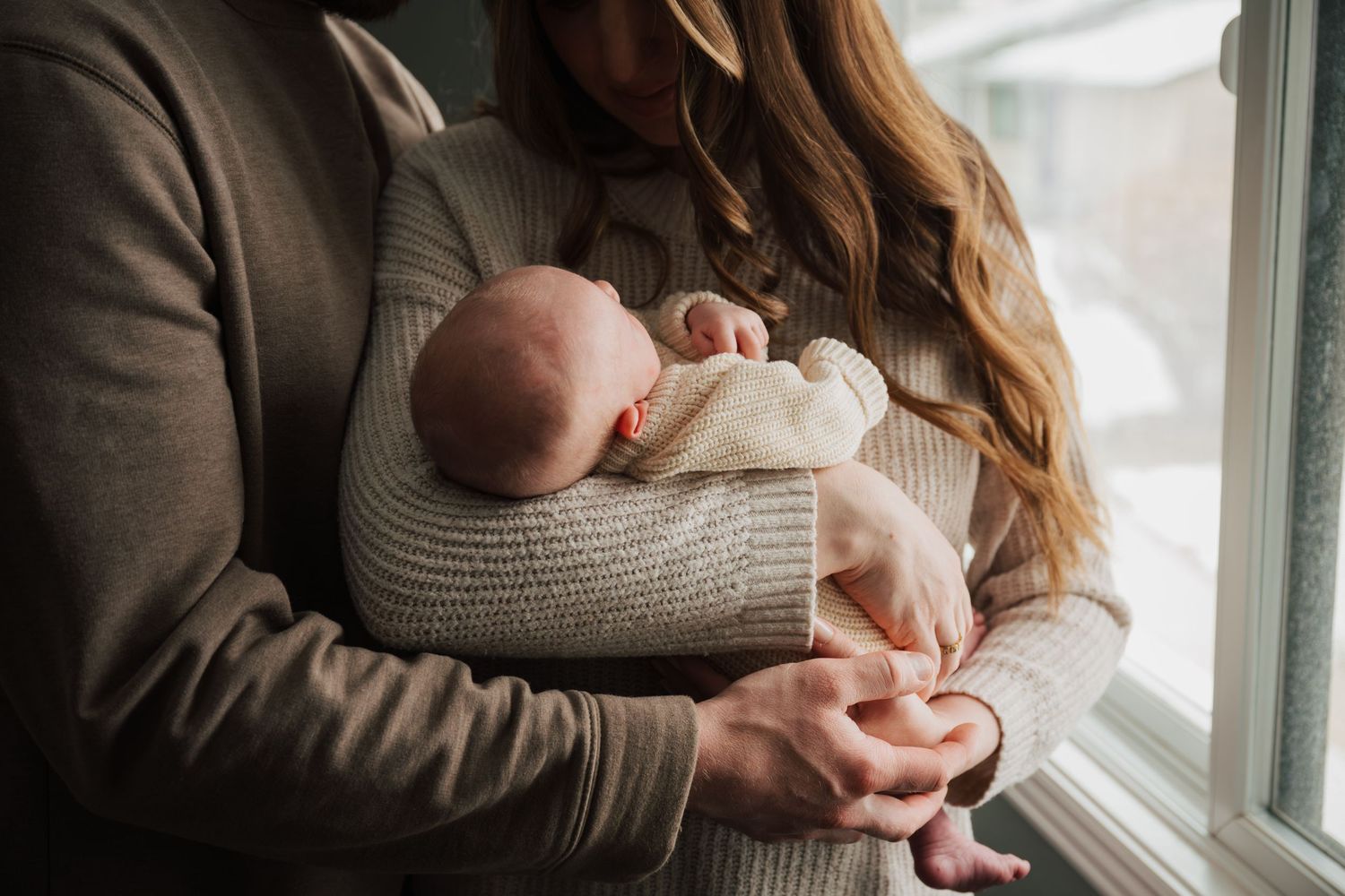Parents sharing intimate moment with newborn by bright window in neutral tones.