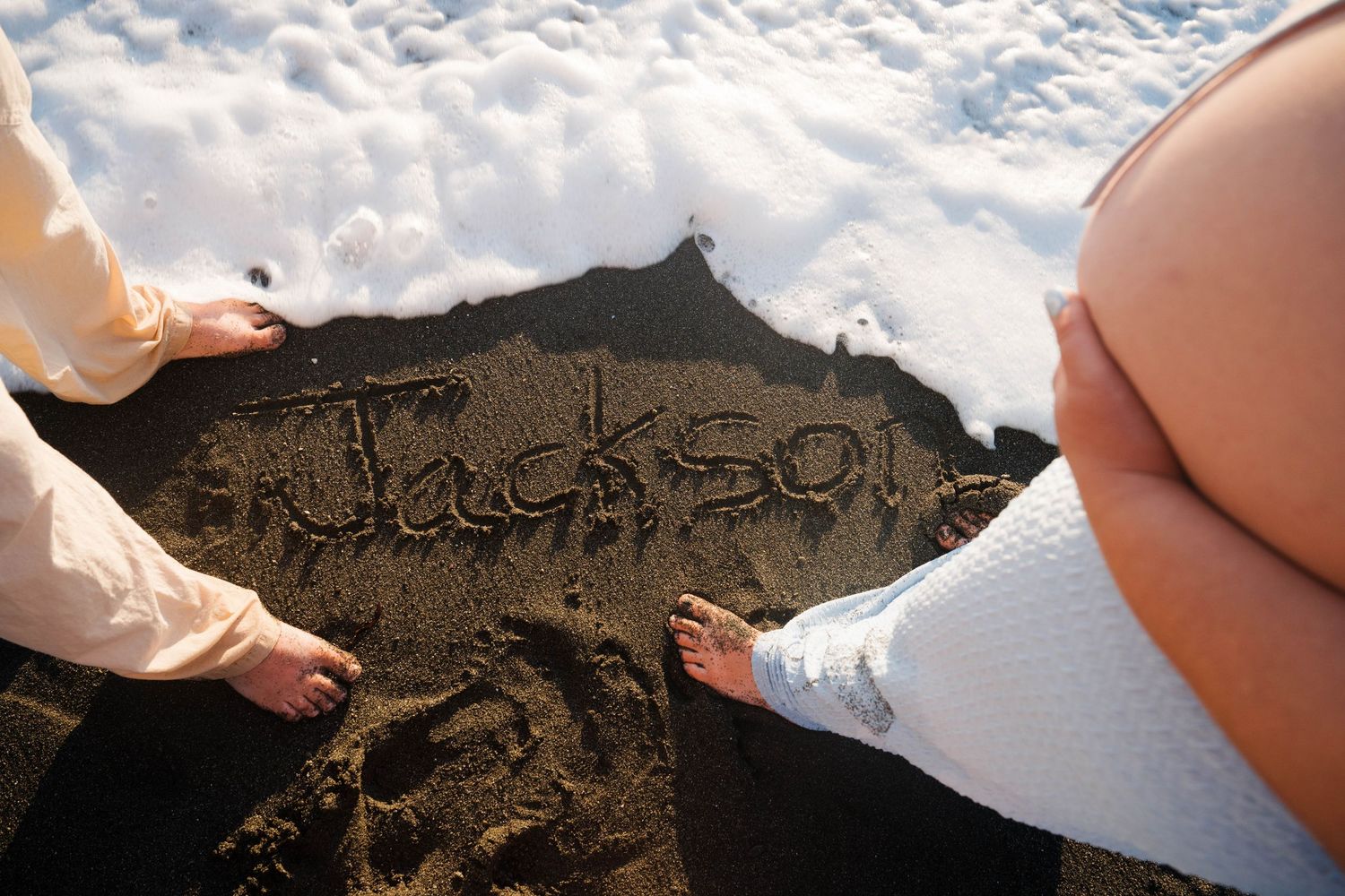 Couple standing barefoot on dark sand beach with name Jackson written in sand near ocean waves.